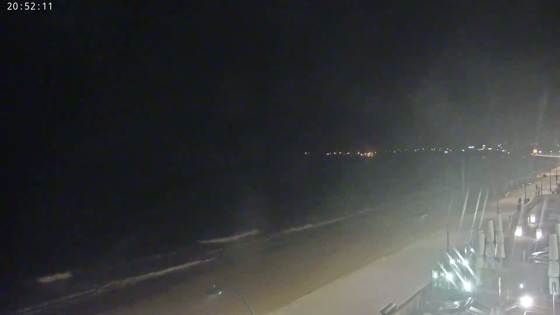 A nighttime beach scene shows gently rolling waves lapping onto a sandy shore, with some lights visible in the distance and folded beach umbrellas on the boardwalk.