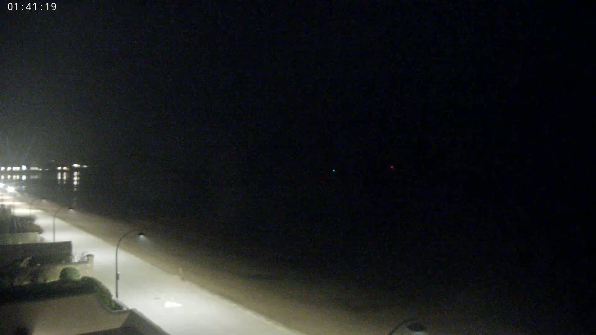 A dark, nighttime view shows a beach, a paved walkway with streetlights, and a calm body of water beyond.