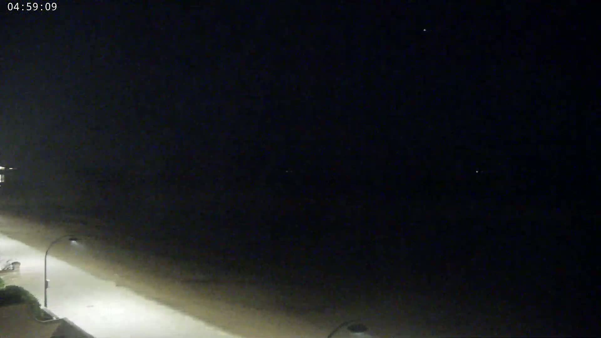 A nighttime beach scene shows a dark ocean under a clear, starlit sky, with a brightly lit paved walkway next to it.