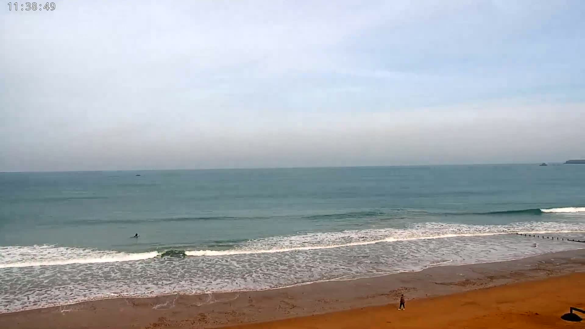 A sandy beach with gentle waves rolls onto the shore under an overcast sky, and a lone person walks along the sand.