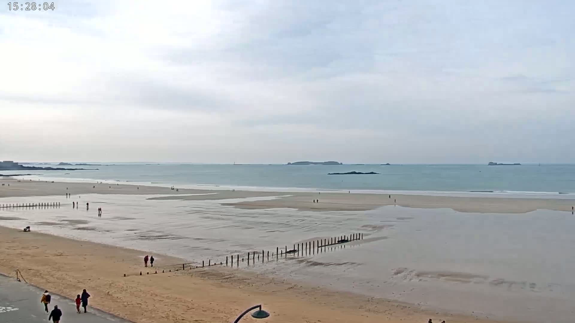 A wide sandy beach with a calm sea under an overcast sky features people walking along the shore and a wooden structure extending into the shallow water.