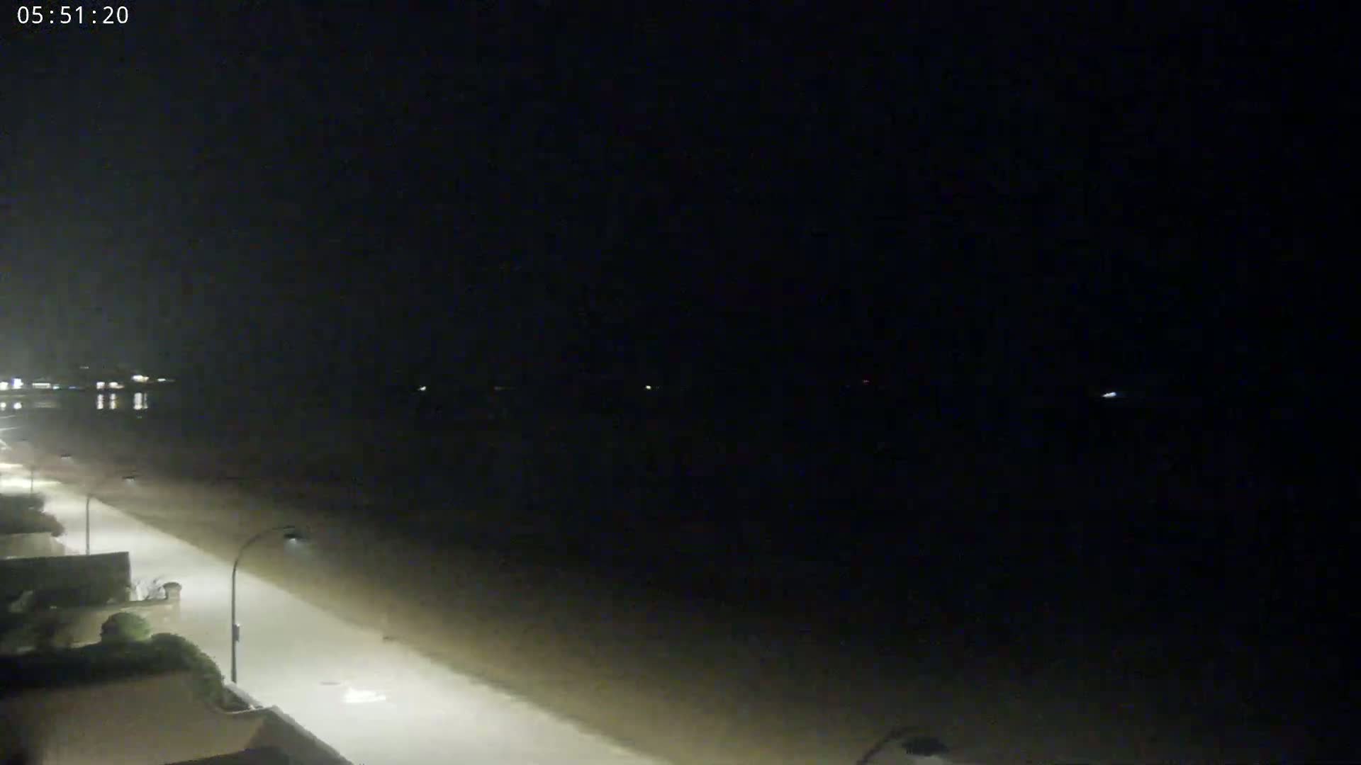 A dark, nighttime view shows a dimly lit beach and road with buildings visible on the left.