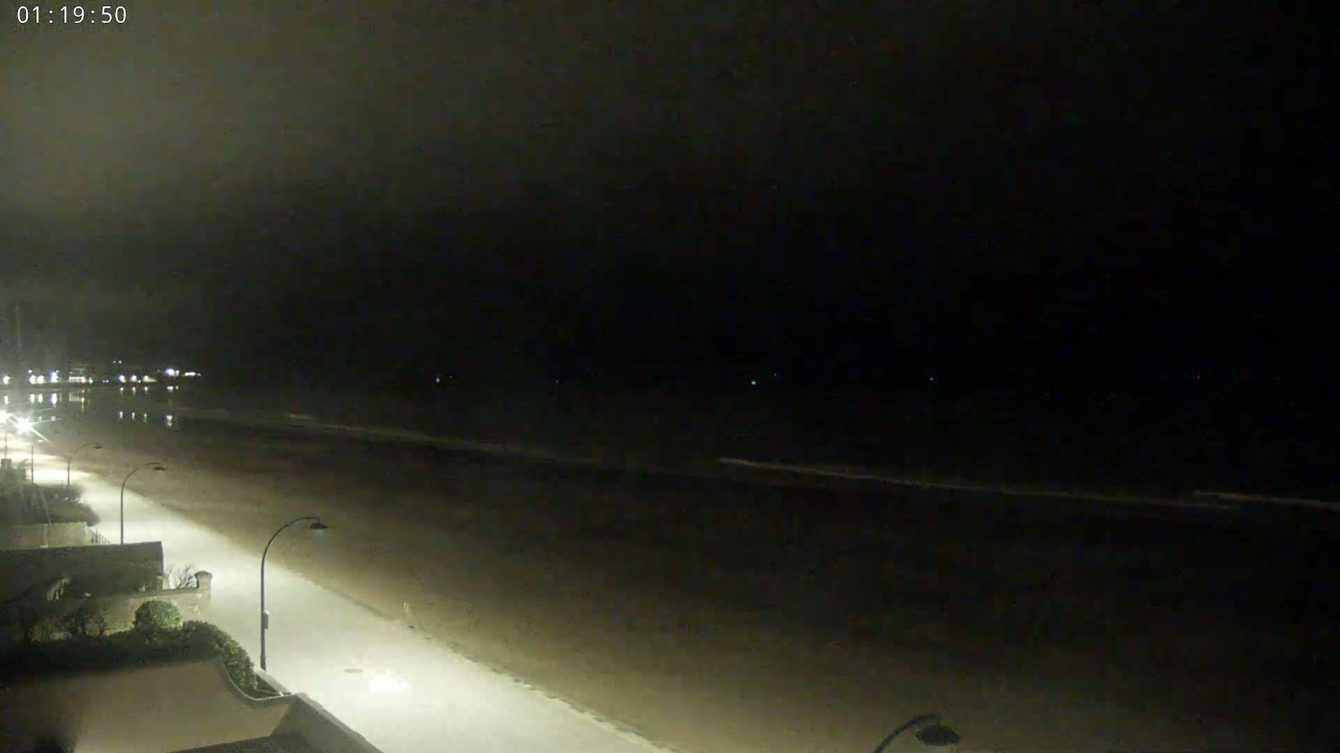 A nighttime view shows a dark beach with calm waves, a paved walkway with streetlights, and buildings in the distance under a cloudy sky.