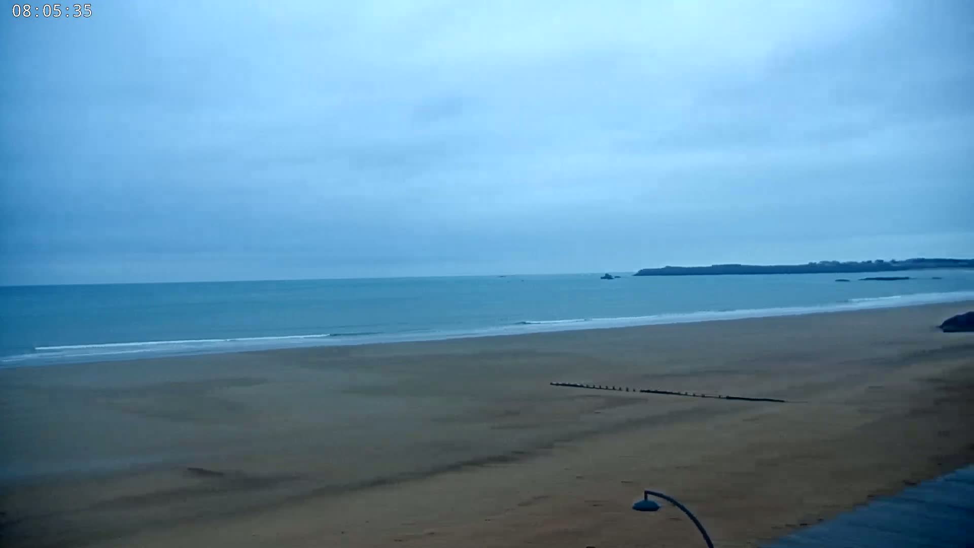 A wide shot shows a sandy beach under an overcast sky, with calm ocean waves lapping at the shore and a distant landmass visible.
