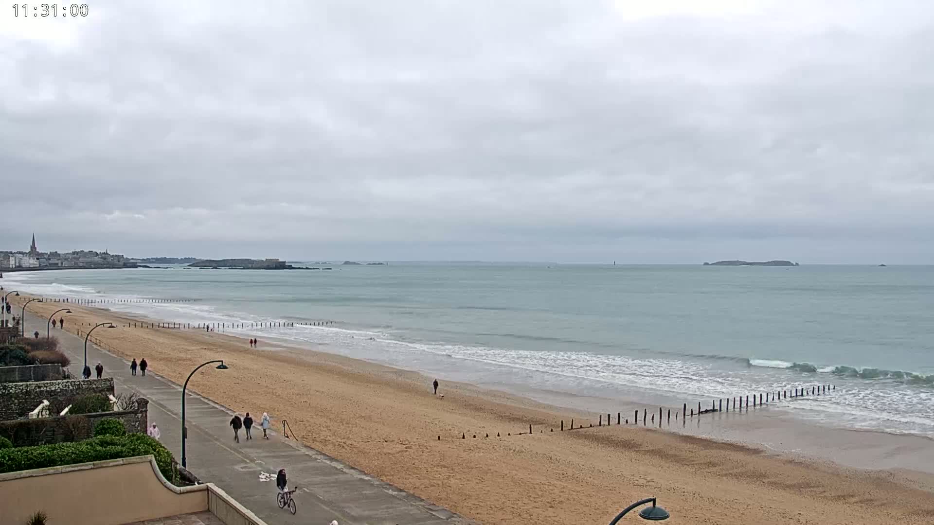 A long sandy beach with gentle waves and a few people walking along the shore under an overcast sky.