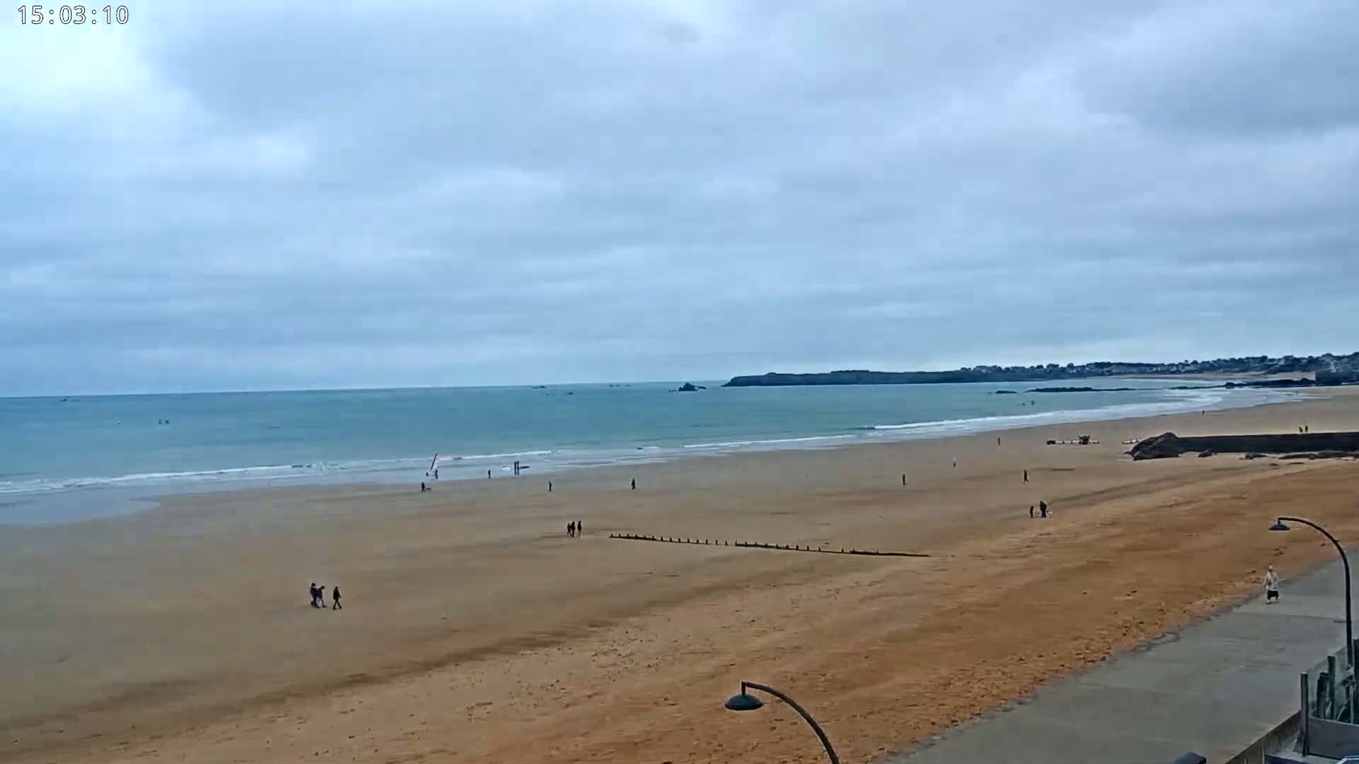 A wide sandy beach stretches to a calm ocean under a cloudy sky, with a few people scattered across the sand.