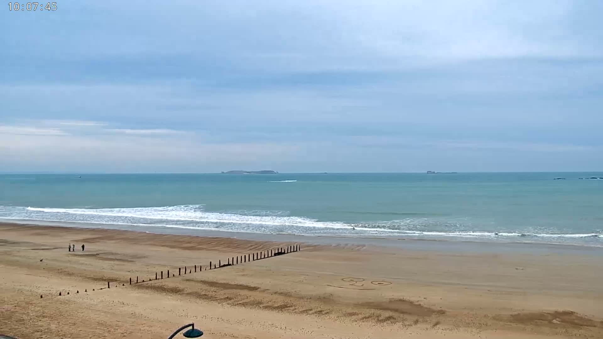 A sandy beach with a few people walking, a wooden post barrier, and a smiley face drawn in the sand stretches out to a calm, light-blue ocean under a mostly cloudy sky.