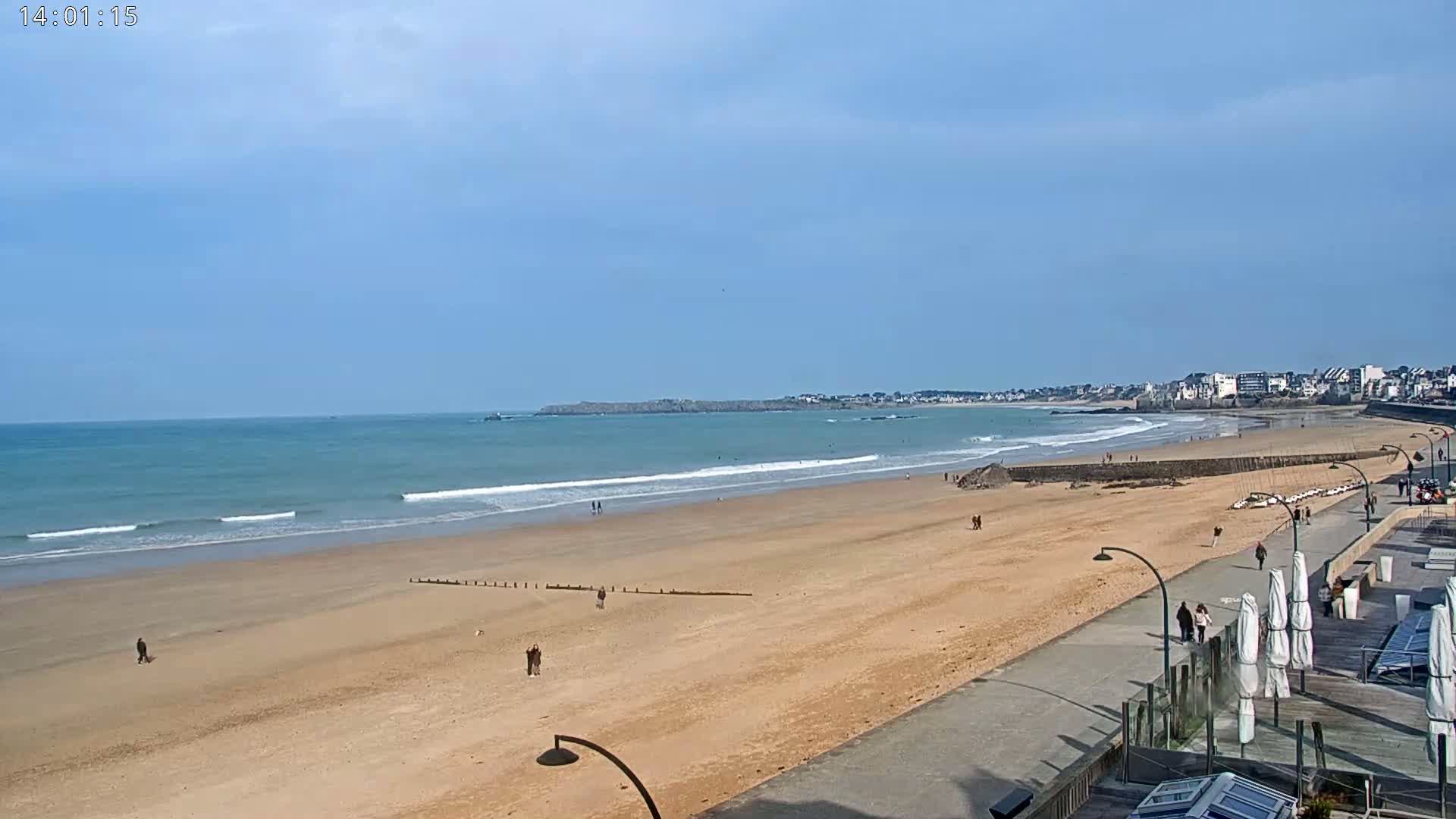 A sandy beach with small waves and a few people stretches out to a town under a mostly clear blue sky.