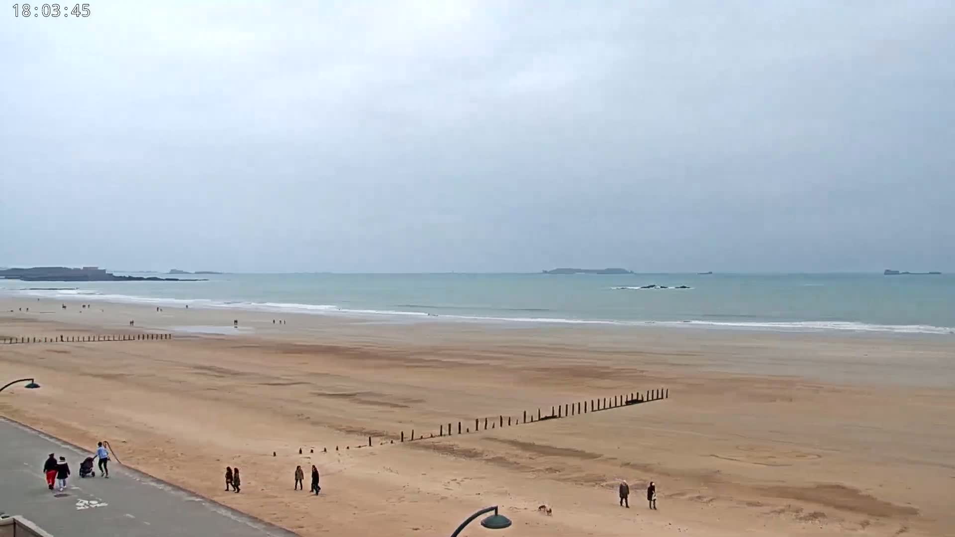 A wide sandy beach stretches to a calm, light-blue ocean under an overcast sky, with several people strolling along the shoreline and a paved walkway.