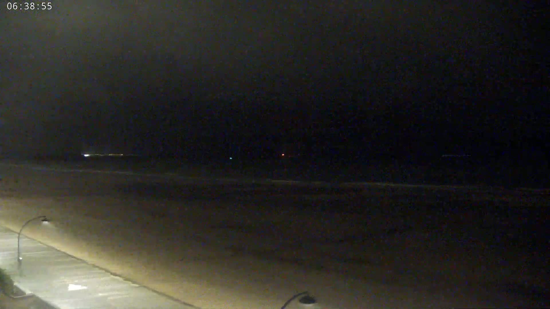 A dark, nighttime view shows a beach with a paved walkway and dimly lit streetlights, under a mostly clear sky.