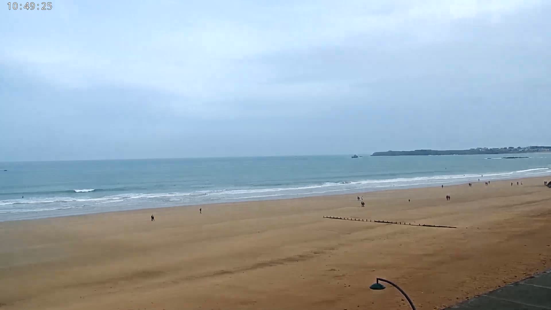 A wide sandy beach stretches to a calm ocean under an overcast sky, with a few people scattered along the shore.