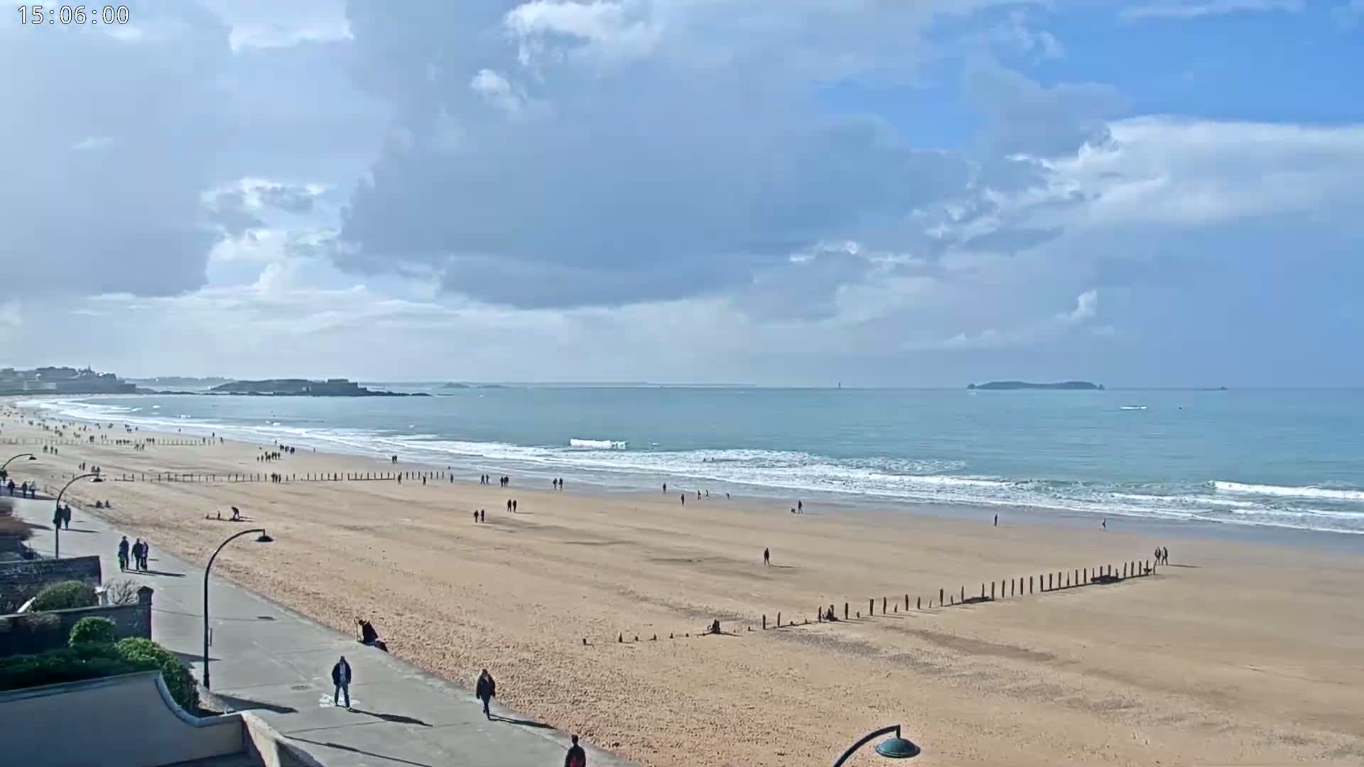A sandy beach with people walking along the shore and a paved walkway, under a partly cloudy sky, with gentle waves breaking on the shore in the distance.