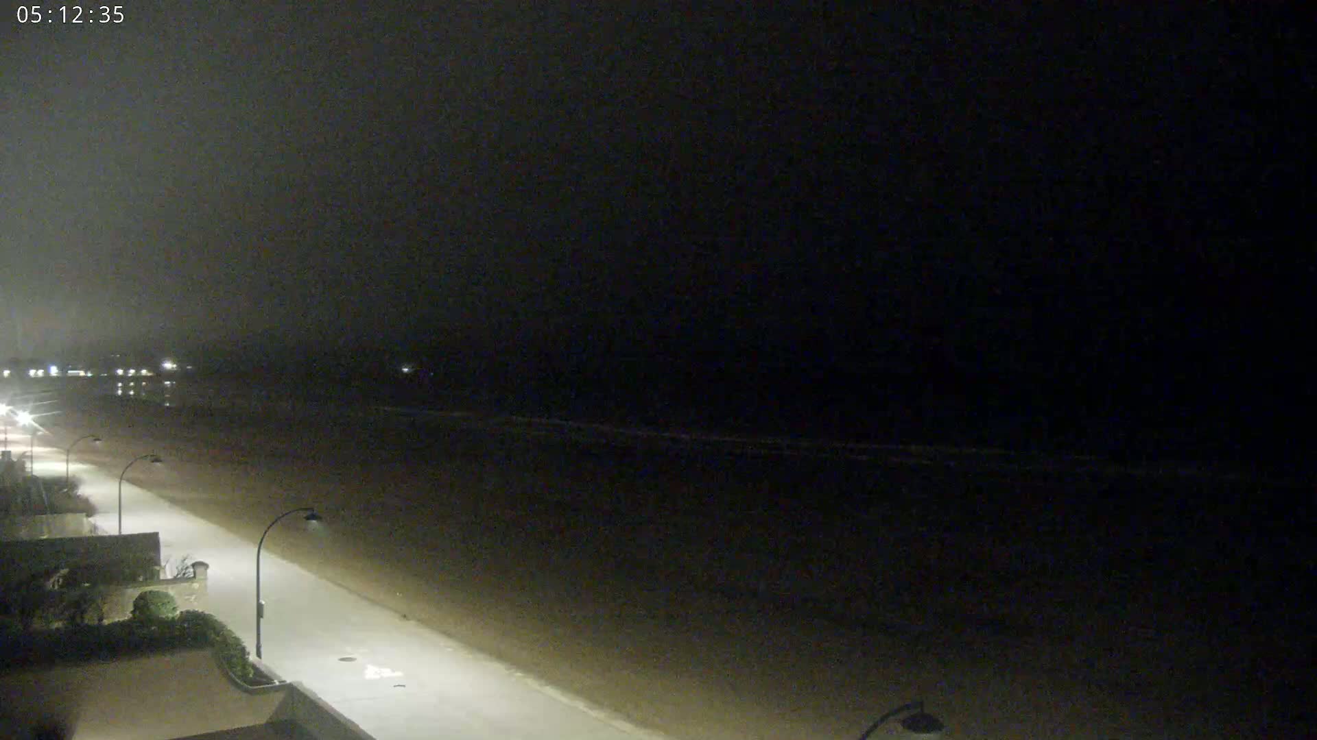 A dark, nighttime view of a beach with a road running alongside it, showing dimly lit buildings in the distance under a mostly cloudy sky.