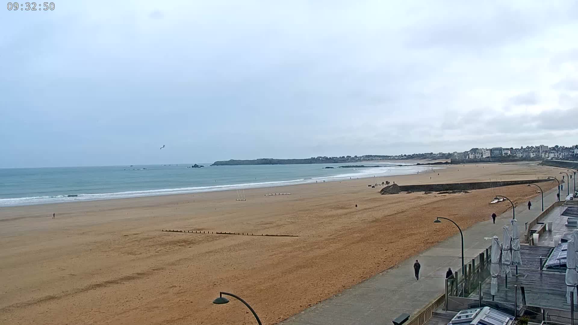 A long sandy beach stretches towards a calm ocean under an overcast sky, with a few people strolling along a paved walkway near buildings in the distance.