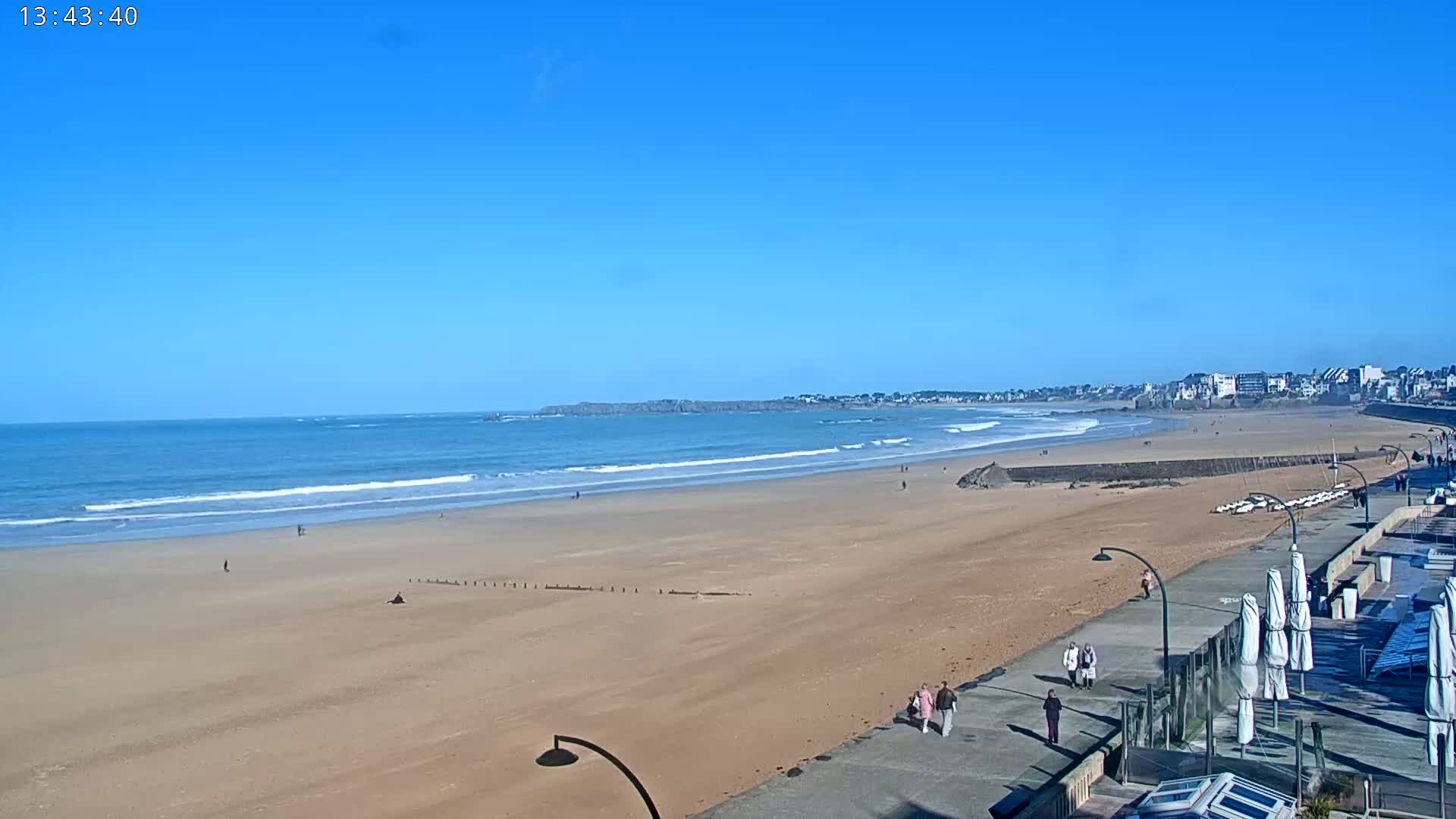 A sandy beach stretches out to a calm ocean under a clear blue sky, with a few people strolling along the boardwalk and a town visible in the distance.