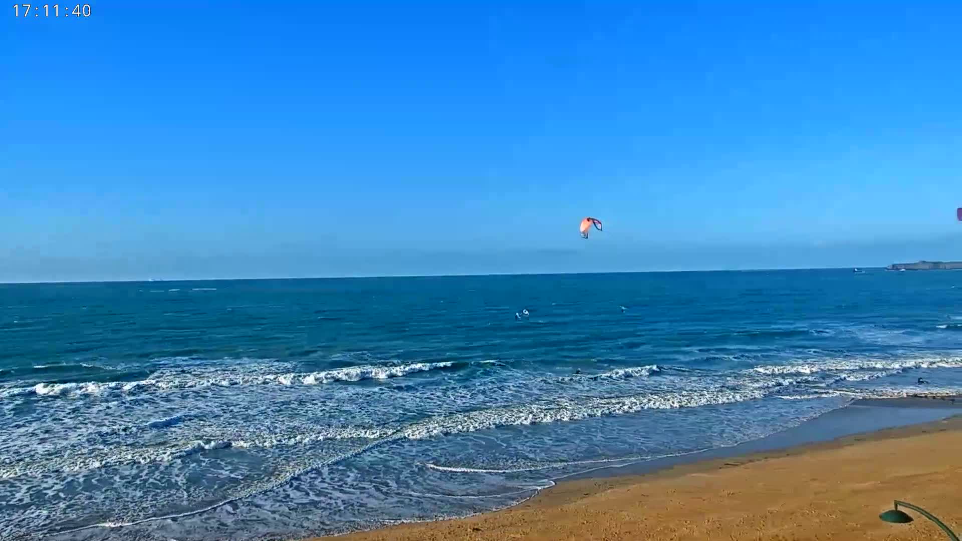 A sandy beach with gentle waves under a clear blue sky, featuring kitesurfers in the ocean.
