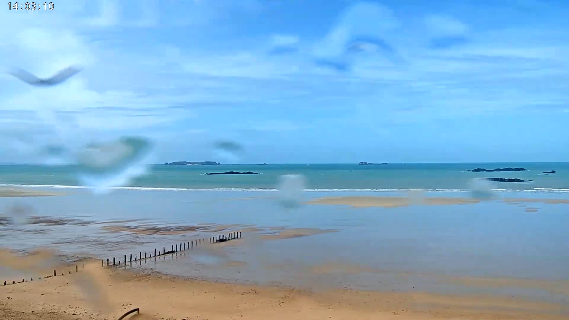 A sandy beach with calm, teal water stretches to distant islands under a mostly clear, light-blue sky, viewed through rain-covered glass.