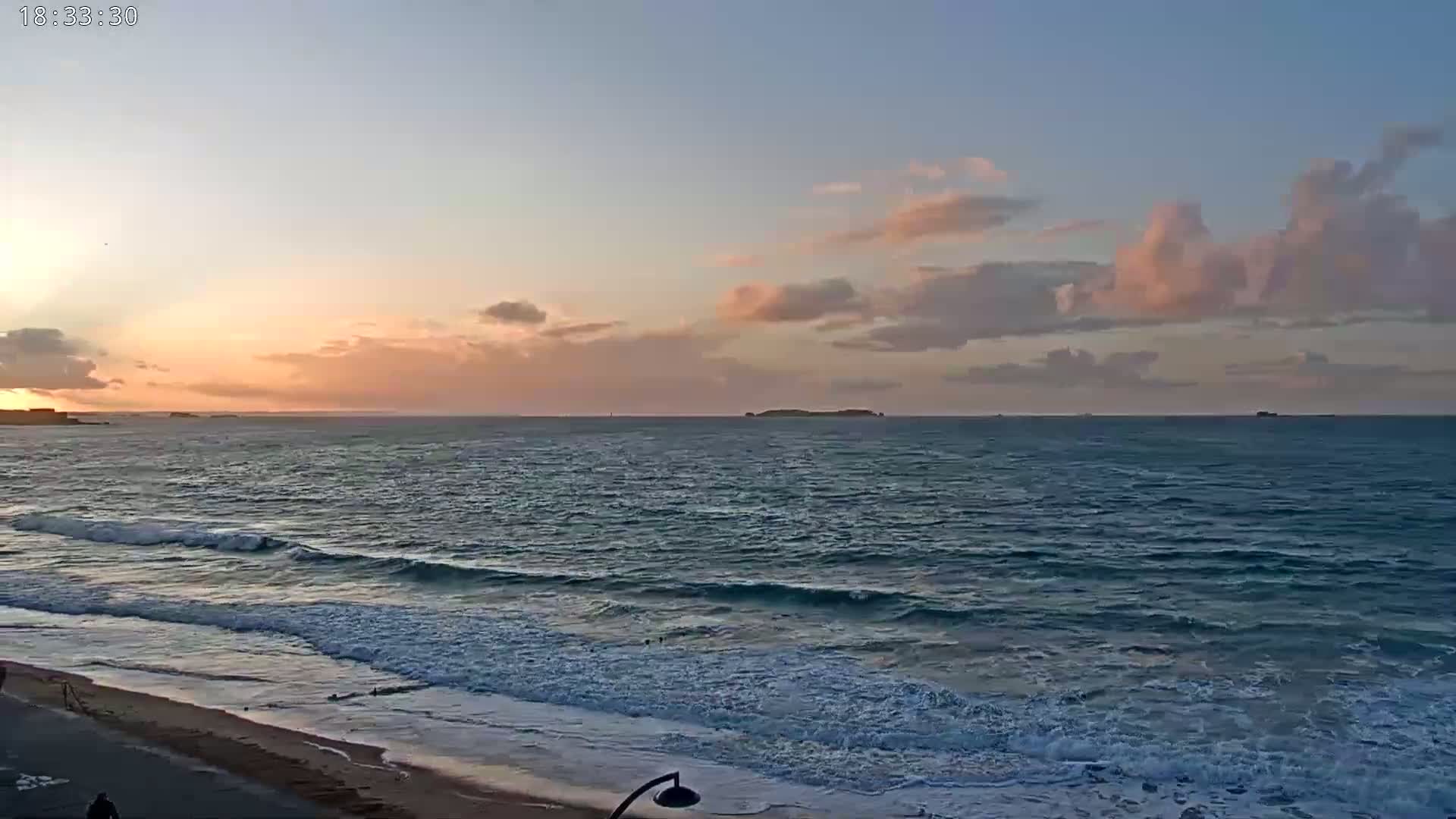 A sandy beach meets a gently waving ocean under a sunset sky with pink and mauve clouds.