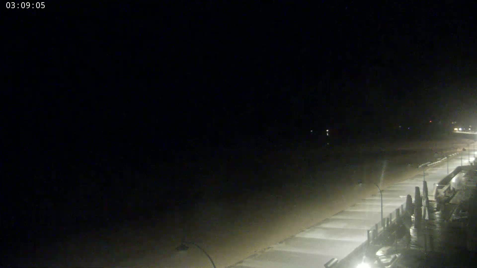 A dark, nighttime view shows a mostly empty beach and boardwalk under a clear sky.