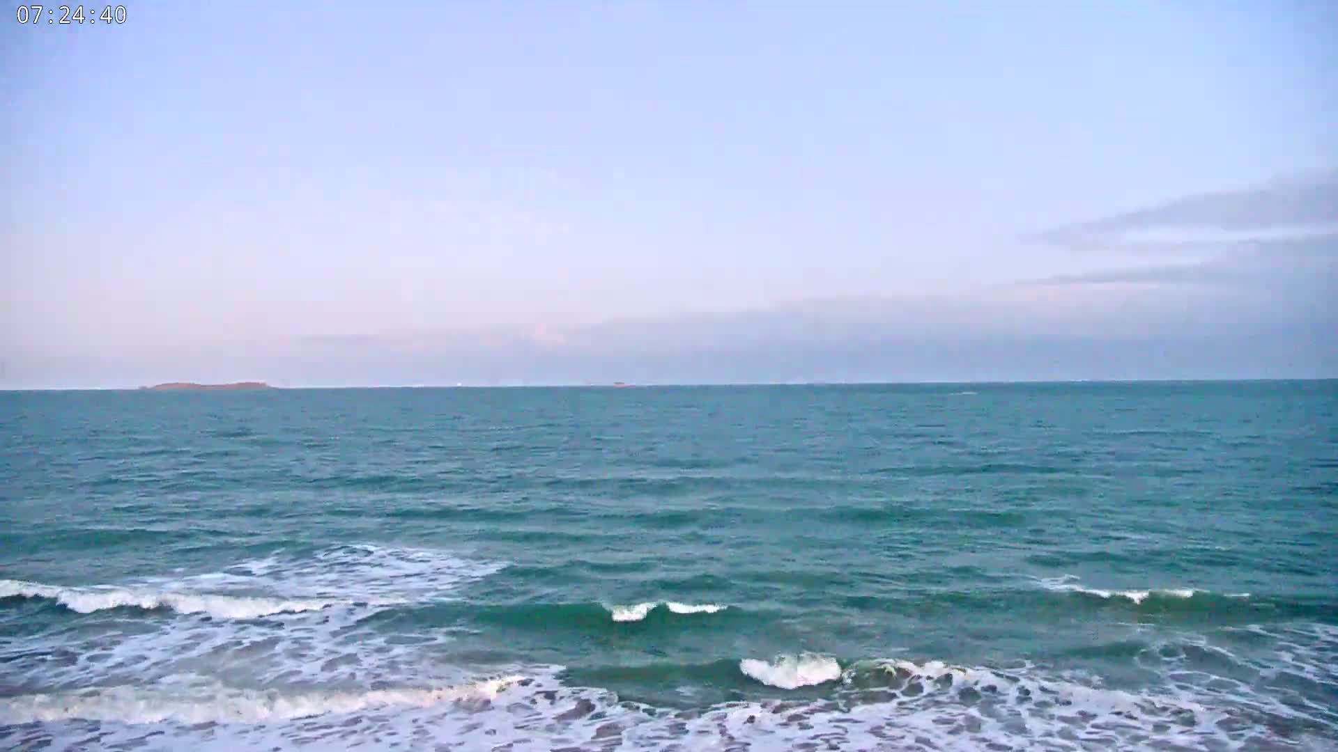 Under a mostly clear sky, teal ocean waves gently lap a sandy shore.