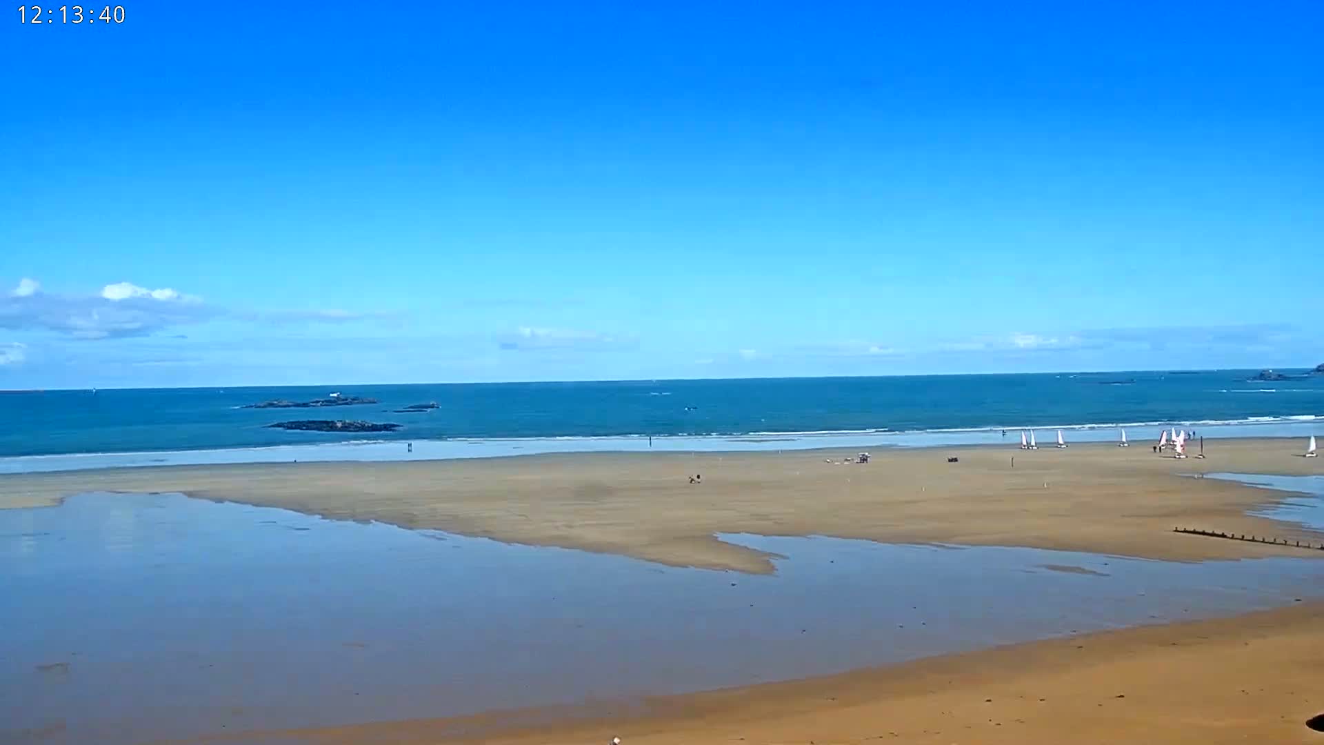 A wide sandy beach with calm blue water under a mostly clear blue sky, with several sailboats in the distance.