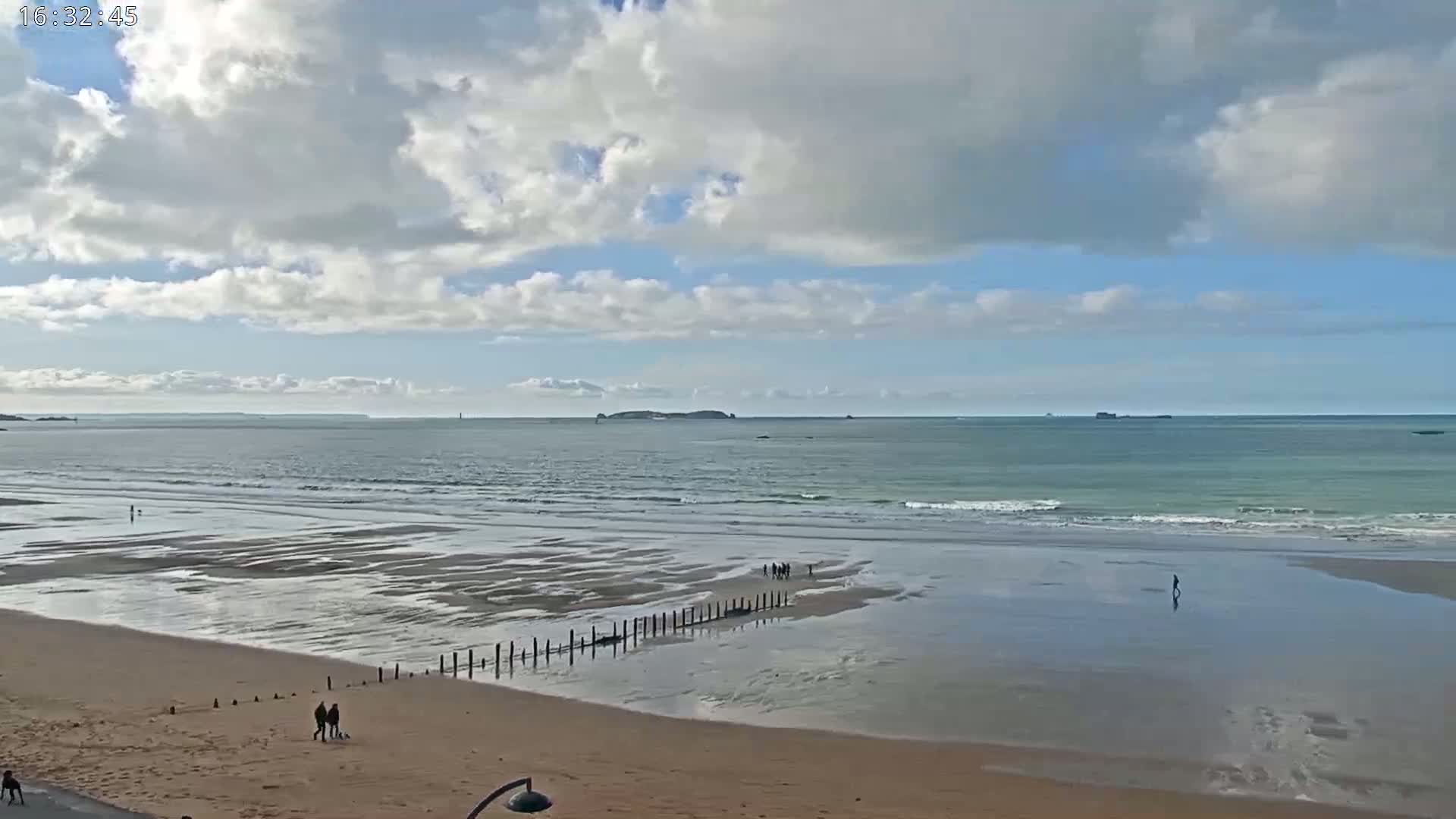 A sandy beach with a calm ocean and a partly cloudy sky shows a few people walking near the water's edge.