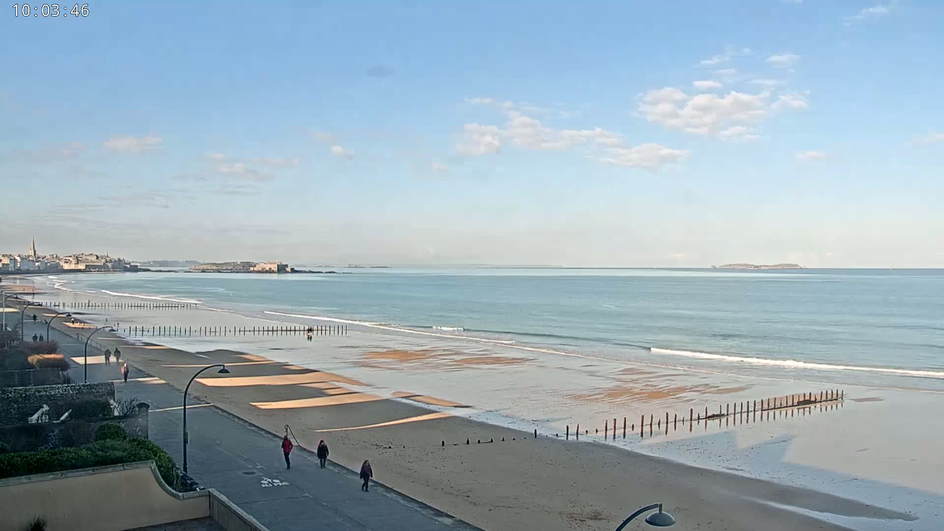 A long sandy beach stretches towards a calm ocean under a mostly clear, bright sky, with a few people walking along the shore path.