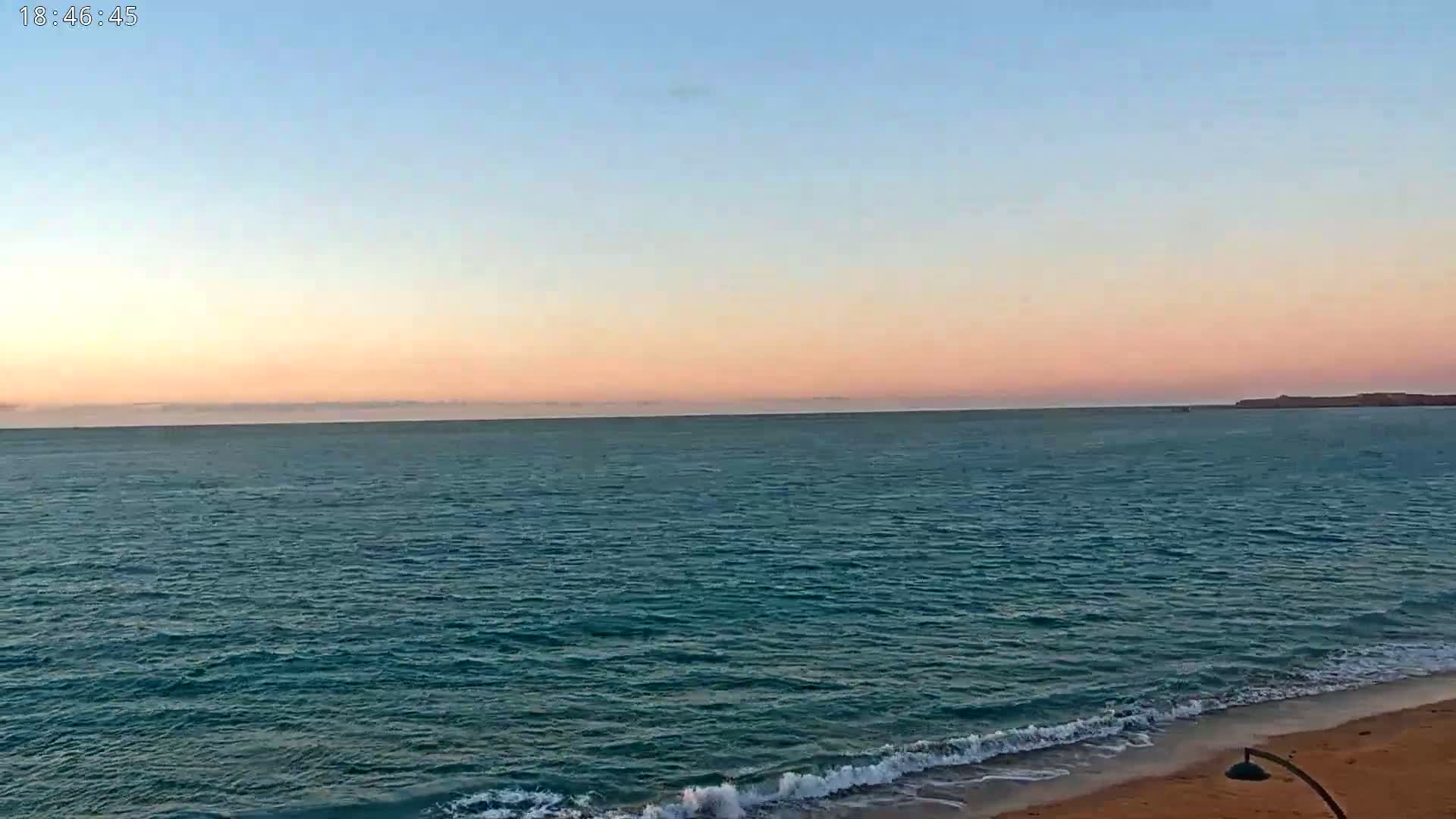 A calm ocean with a sandy beach at sunset under a pale pink and blue sky.
