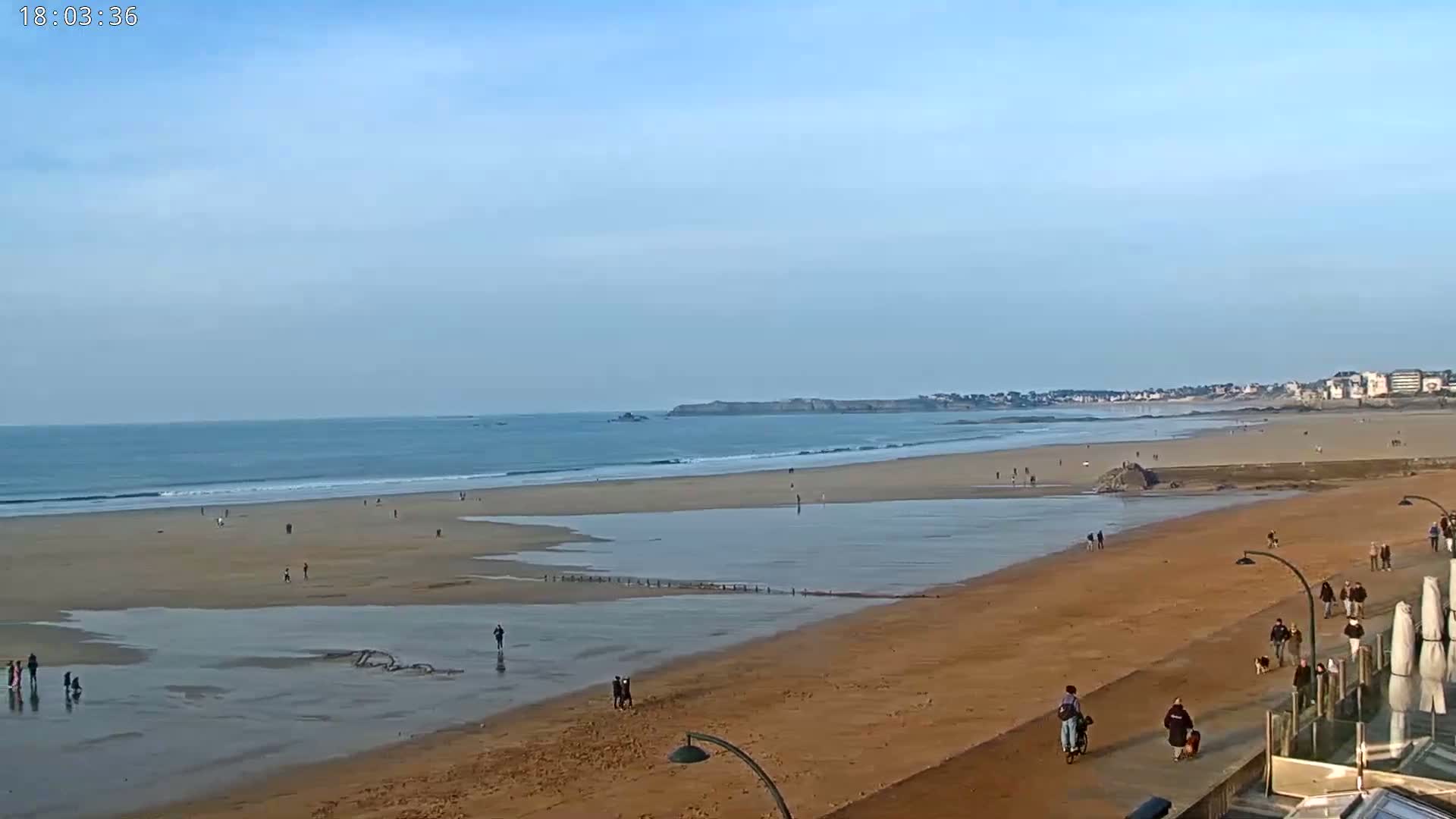 A wide sandy beach with scattered people stretches along a calm ocean under a clear blue sky.