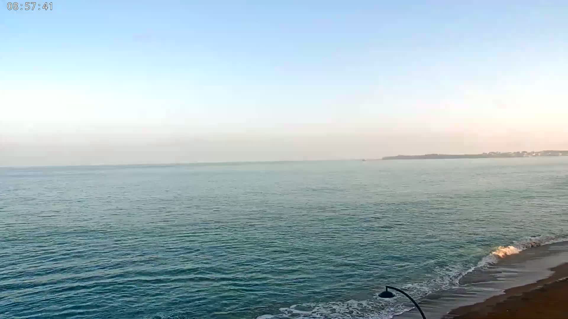 A calm, light-blue ocean meets a sandy beach under a clear, pale-blue sky.