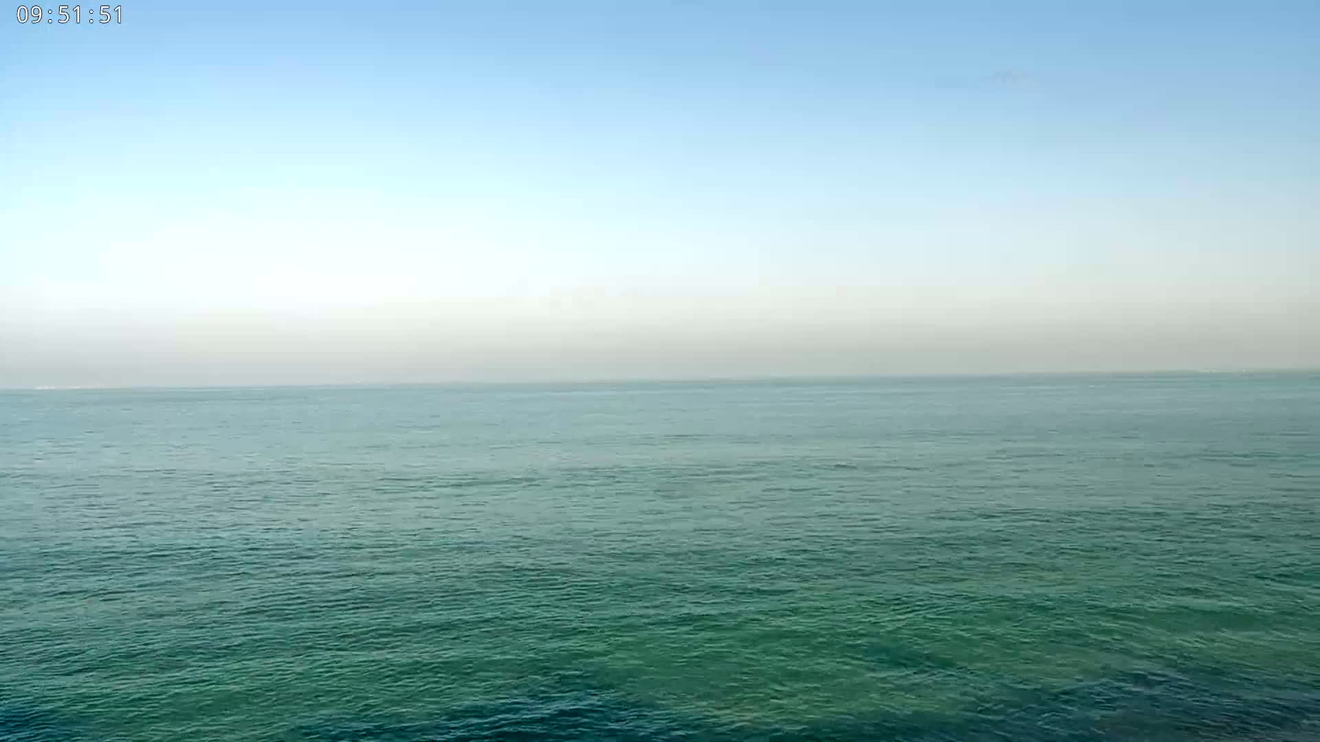 A calm, teal ocean stretches to a pale, hazy horizon under a clear blue sky.