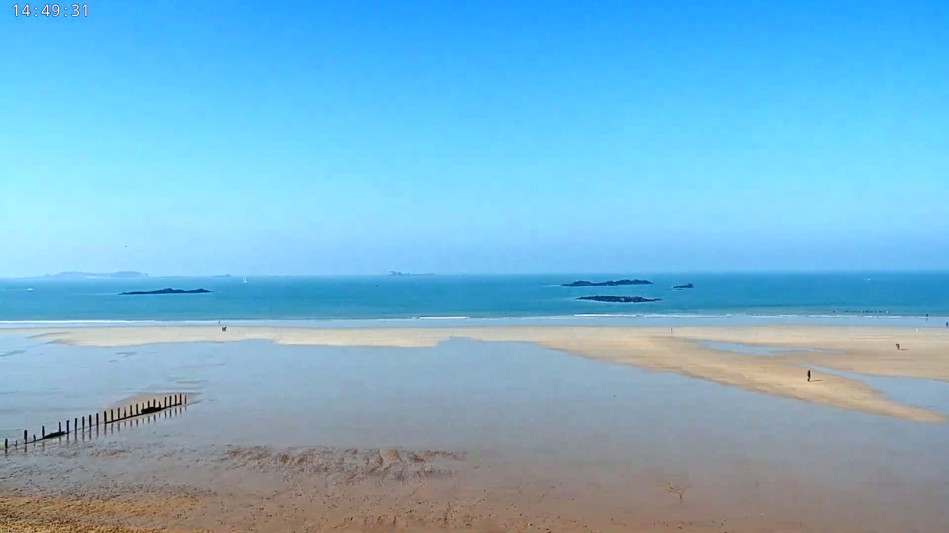 A sandy beach stretches out to a calm, blue ocean under a clear, bright blue sky, with a few people visible on the shore and some distant islands.