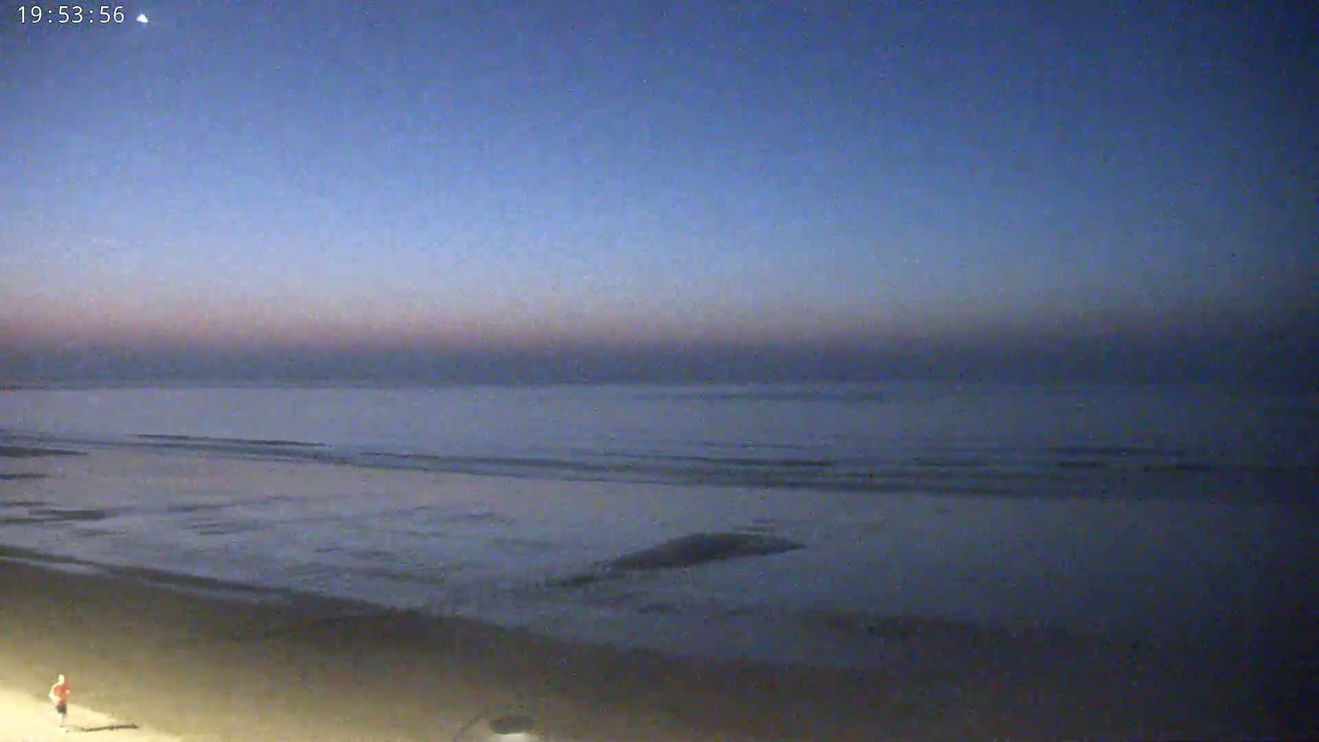 A person walks on a sandy beach near a calm, dark-blue ocean under a twilight sky.