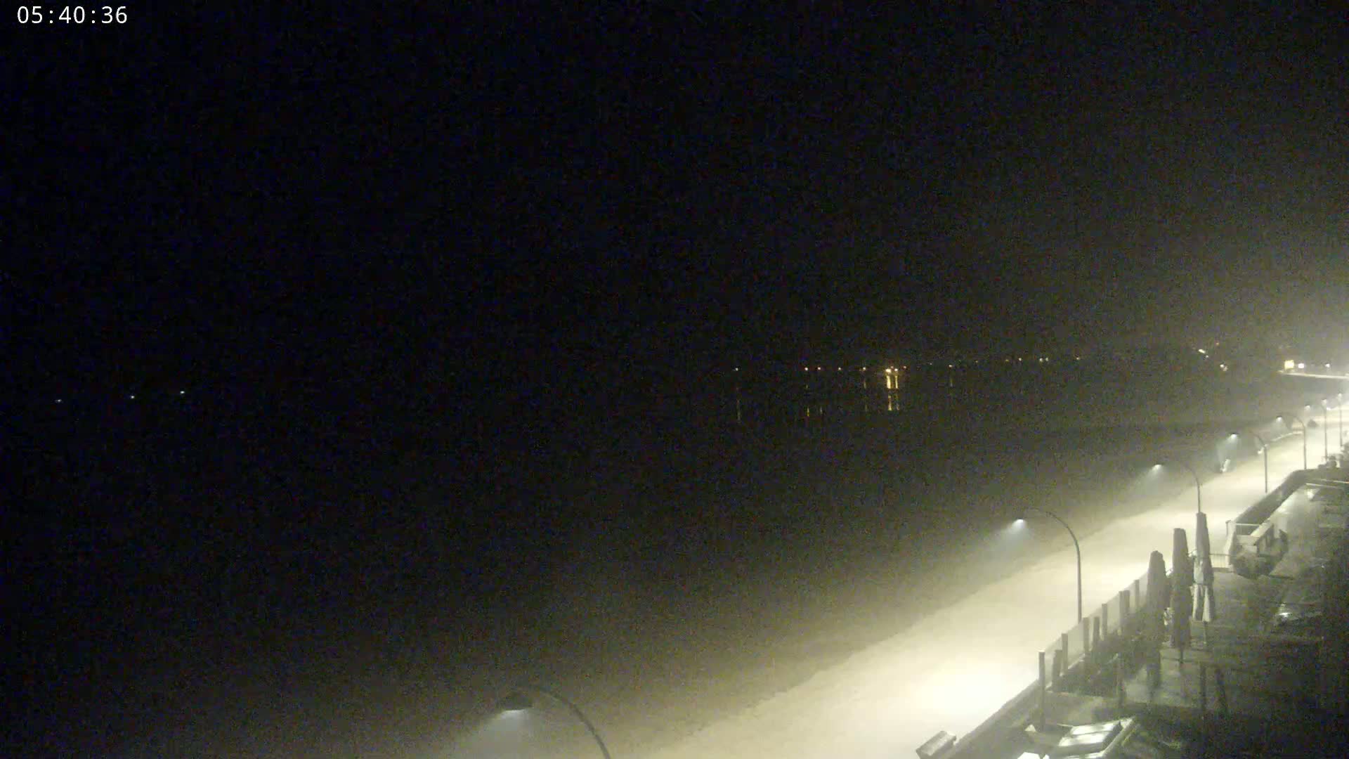 A hazy night scene shows a paved waterfront area with lampposts, a body of water, and faint lights across the water in the distance.