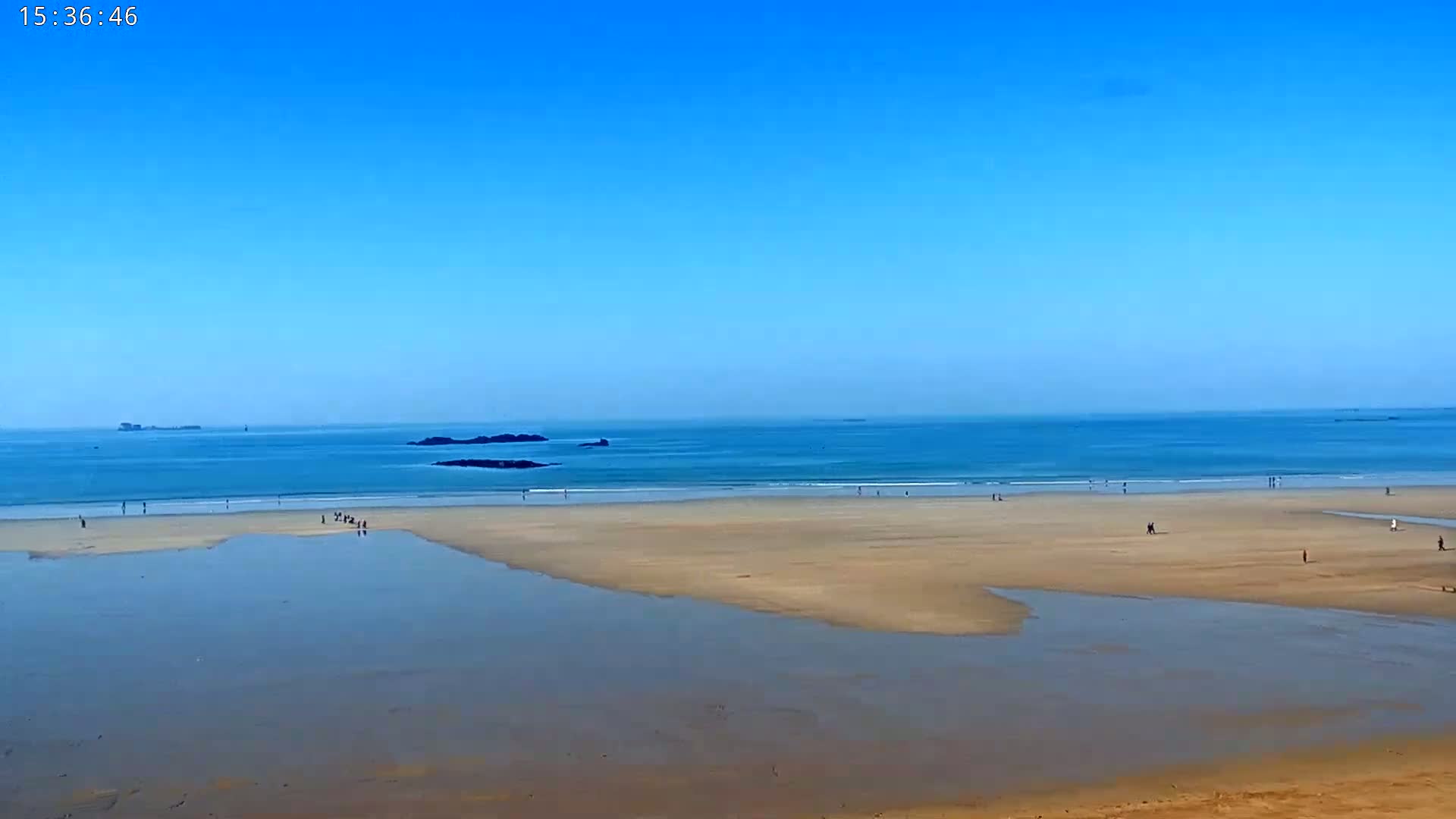 A sandy beach with scattered people stretches out to a calm blue ocean under a clear blue sky.
