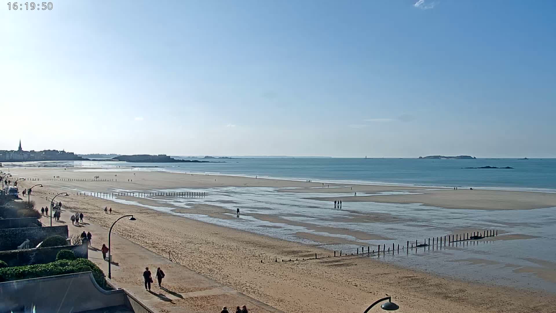 A sandy beach with scattered people stretches toward a calm ocean under a clear blue sky, with a town visible in the distance.