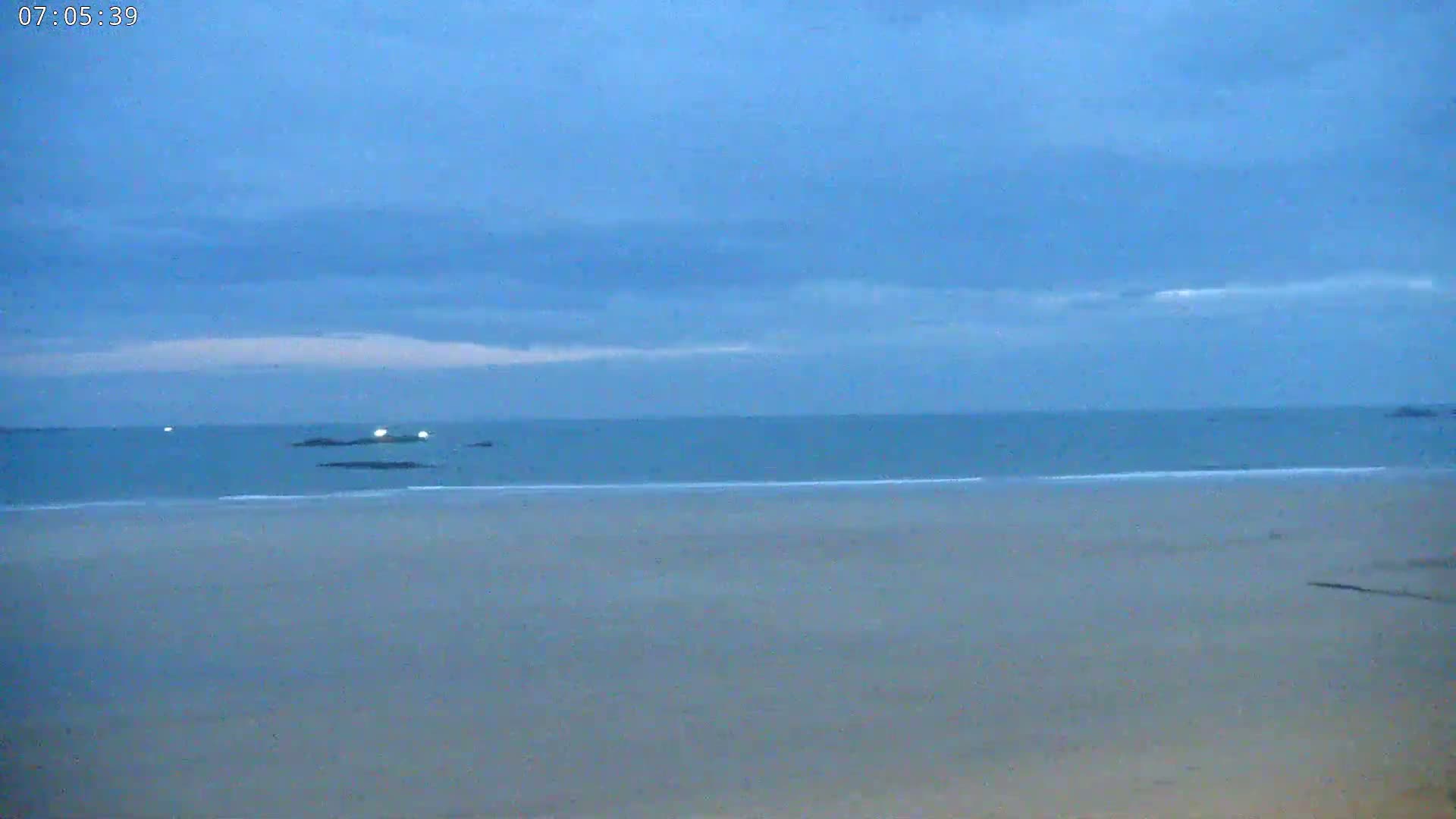 A mostly-clear, blue sky over a calm, light-blue ocean and a wide, flat sandy beach.
