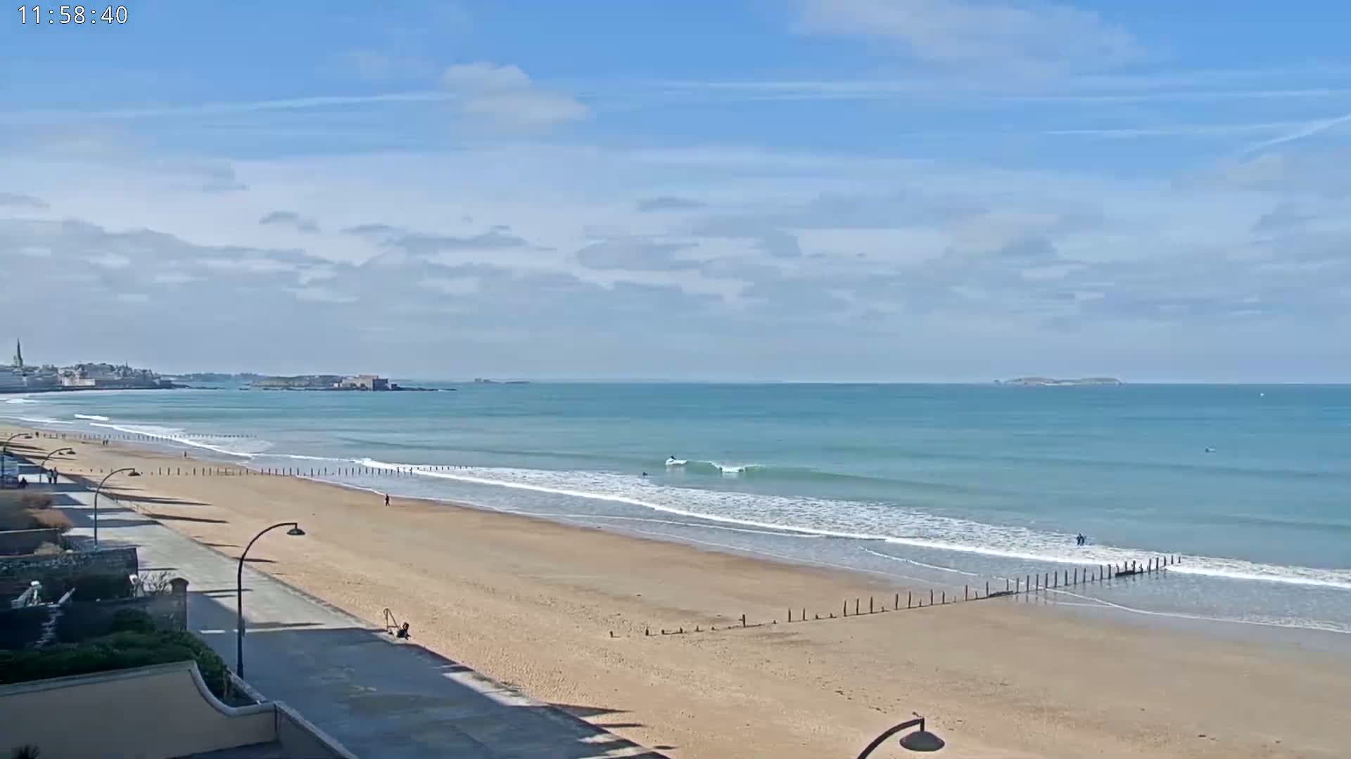 A sandy beach with a few people on it and gentle waves rolling onto the shore under a mostly sunny sky.