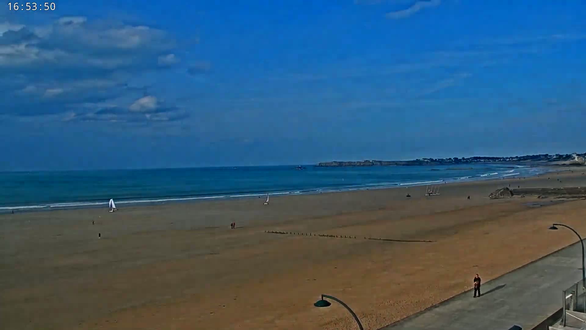 A mostly sunny day at a wide sandy beach with a few people and small sailboats in the water, and a person walking along a paved path at the beach's edge.