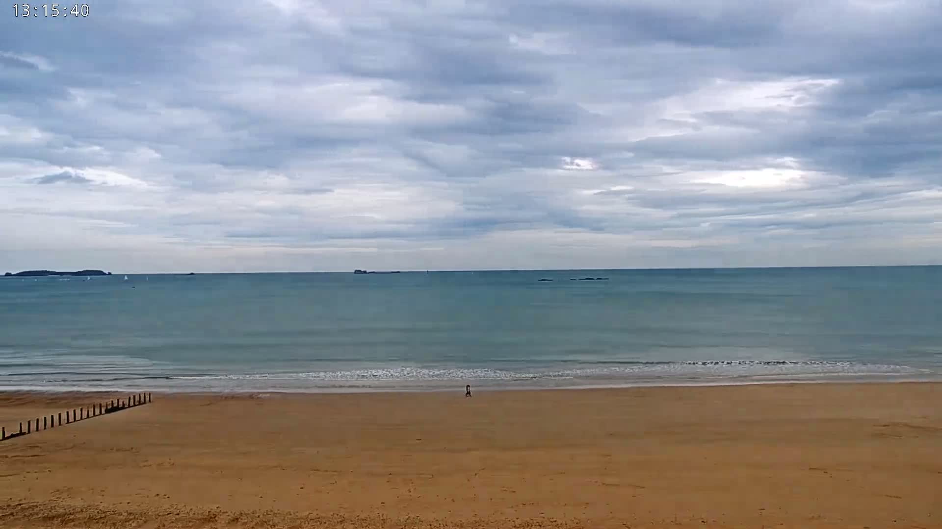 A sandy beach stretches to a calm sea under a mostly cloudy sky, with a single person visible in the distance.