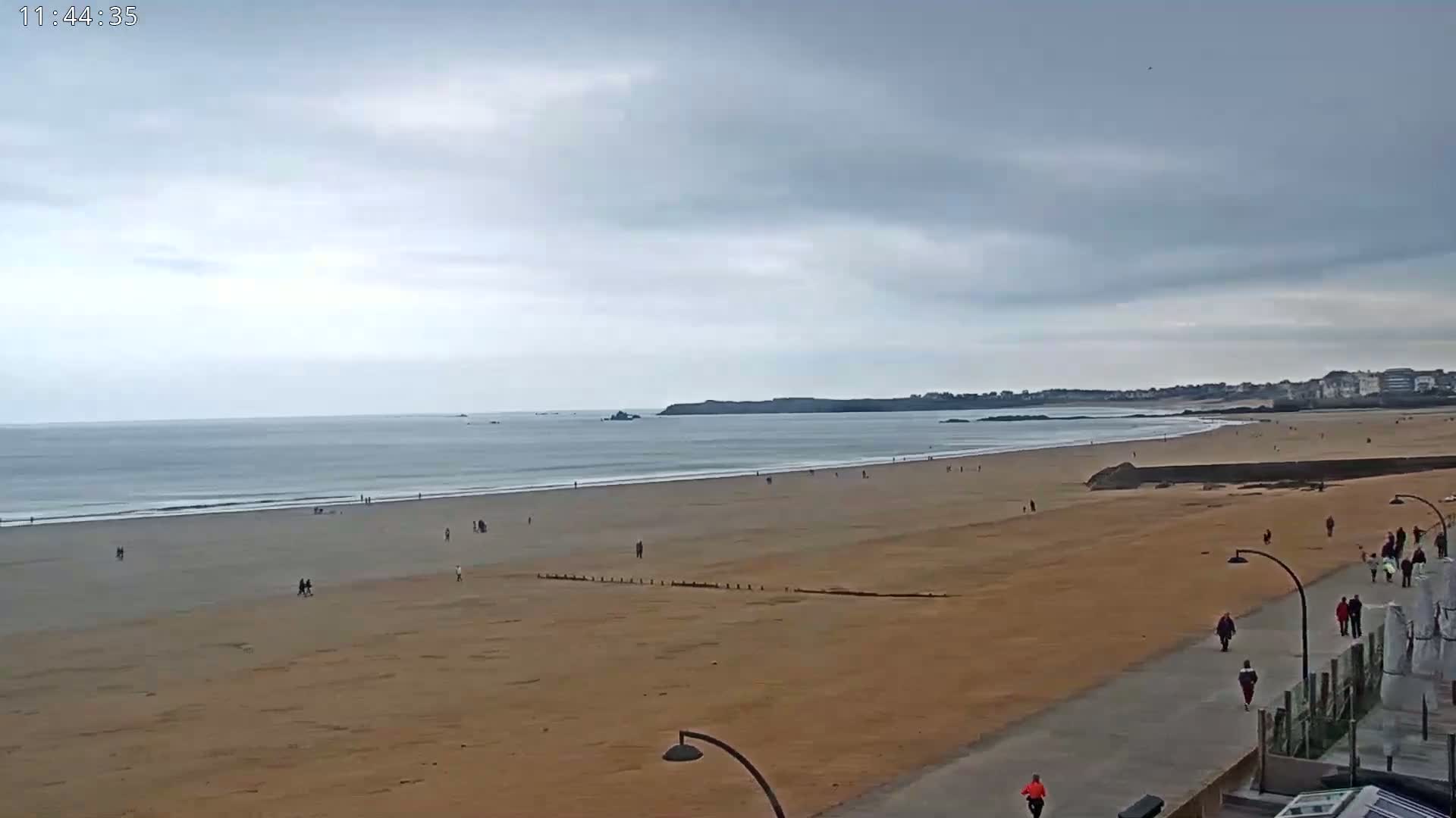 A wide sandy beach stretches to a calm ocean under an overcast sky, with a few people walking on the sand and boardwalk.