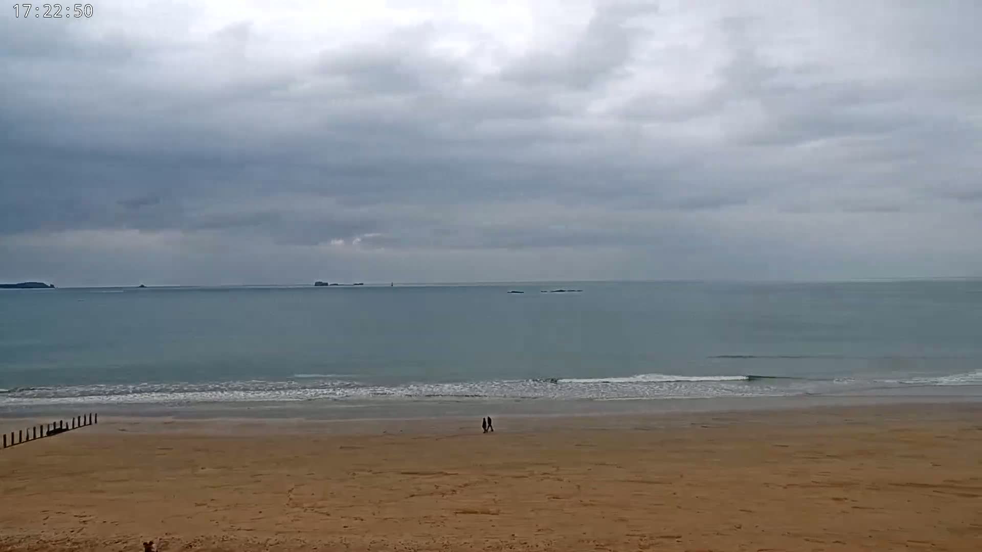 Two people walk on a sandy beach under a cloudy sky, toward a calm ocean with small waves.