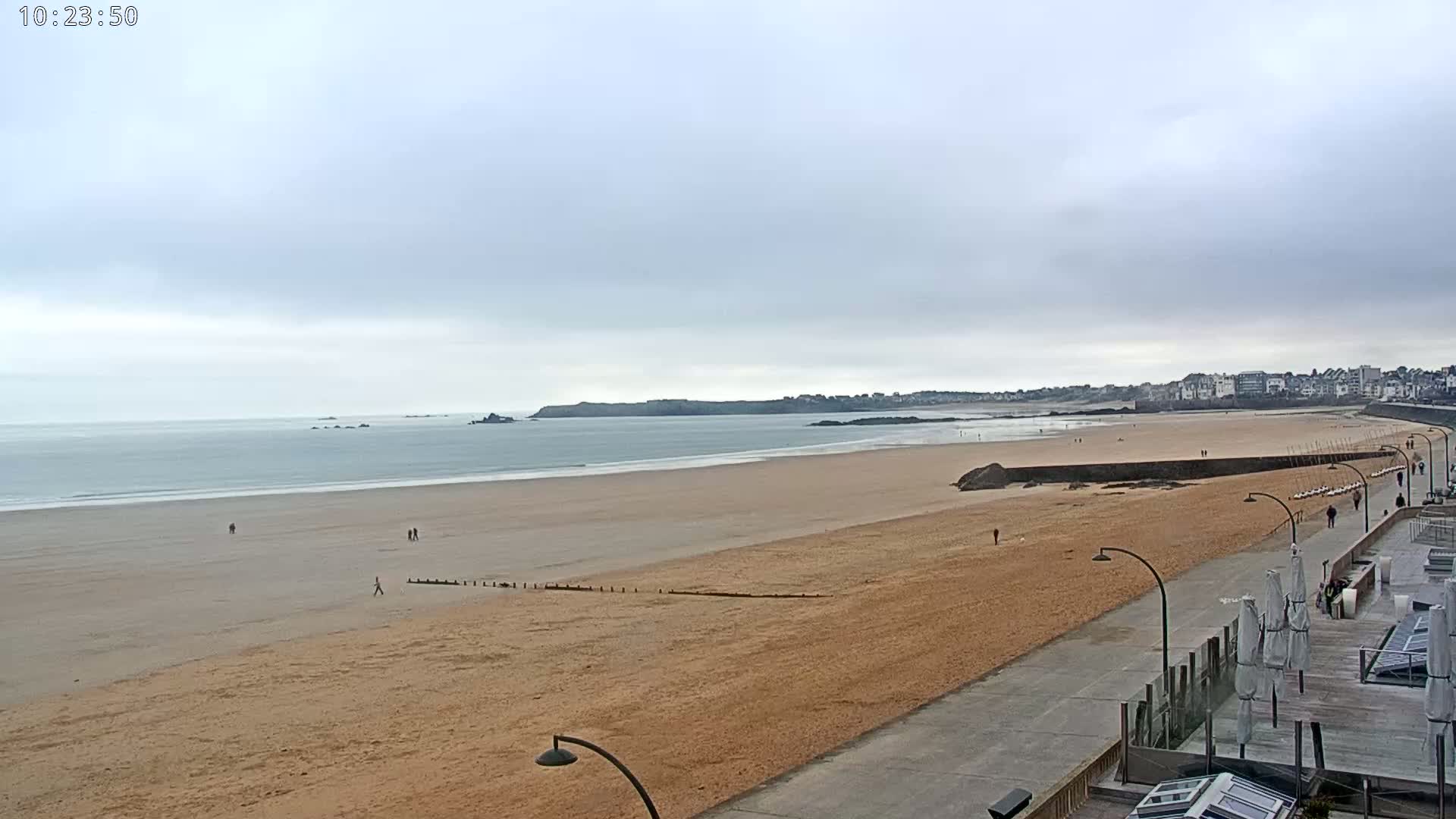A long sandy beach stretches toward a calm ocean under an overcast sky, with a boardwalk and buildings visible in the distance.