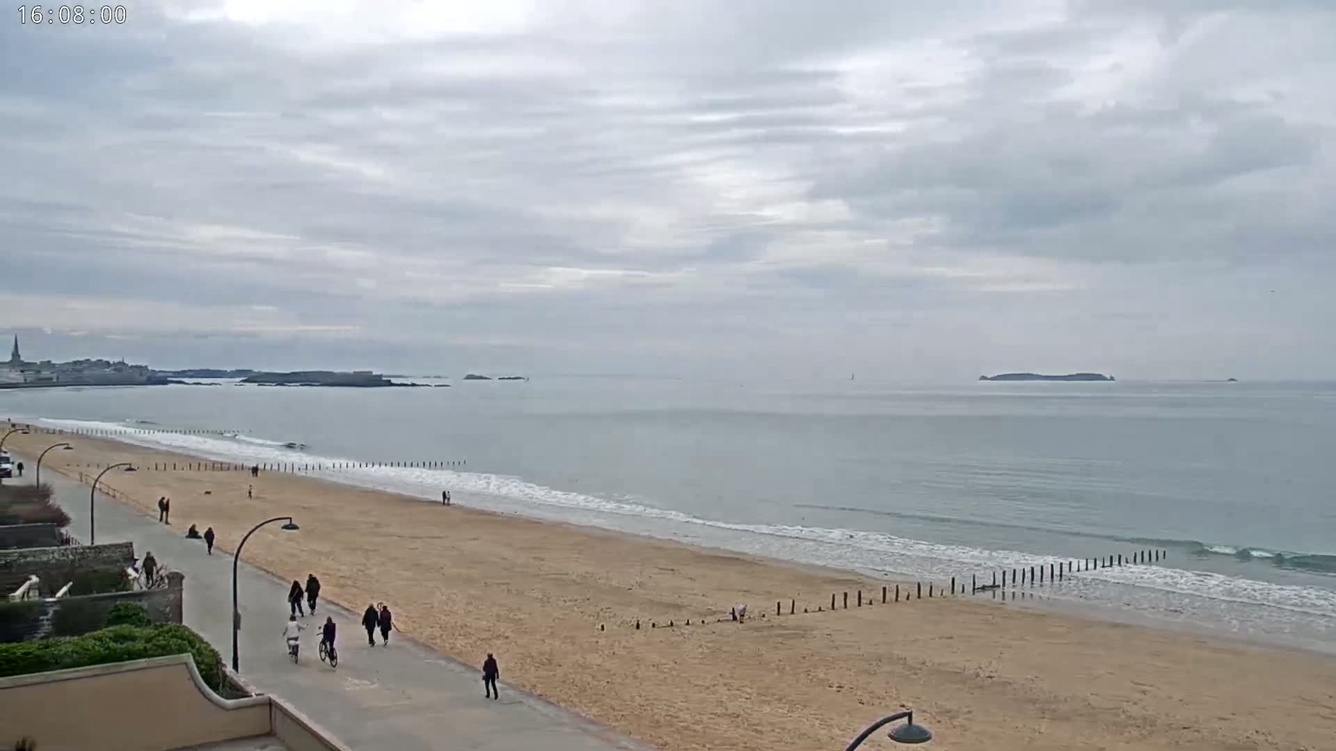 A sandy beach with people walking along a paved path beside it, gentle waves lapping the shore under a mostly cloudy sky.