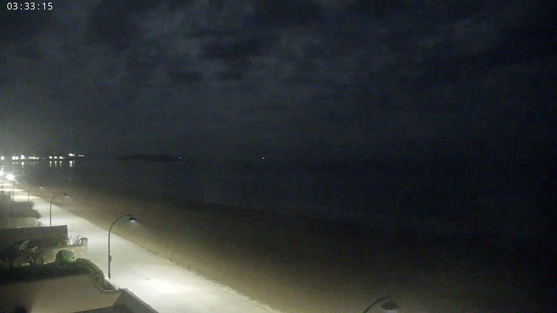 A nighttime view of a mostly empty beach, with a streetlight-lit path leading to a calm, dark ocean under a cloudy sky.