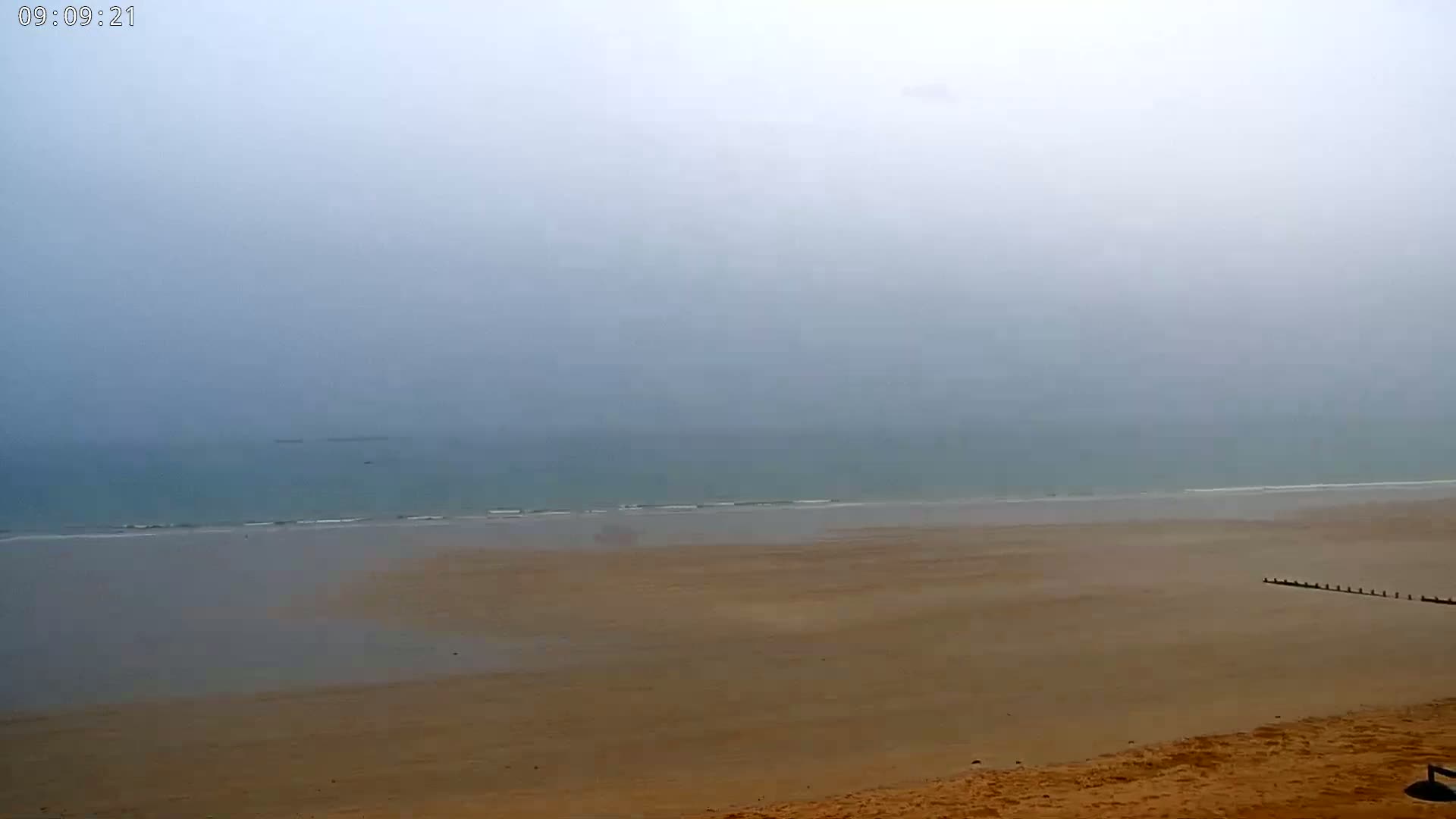 A wide, sandy beach stretches to a calm, foggy ocean under a gray sky.
