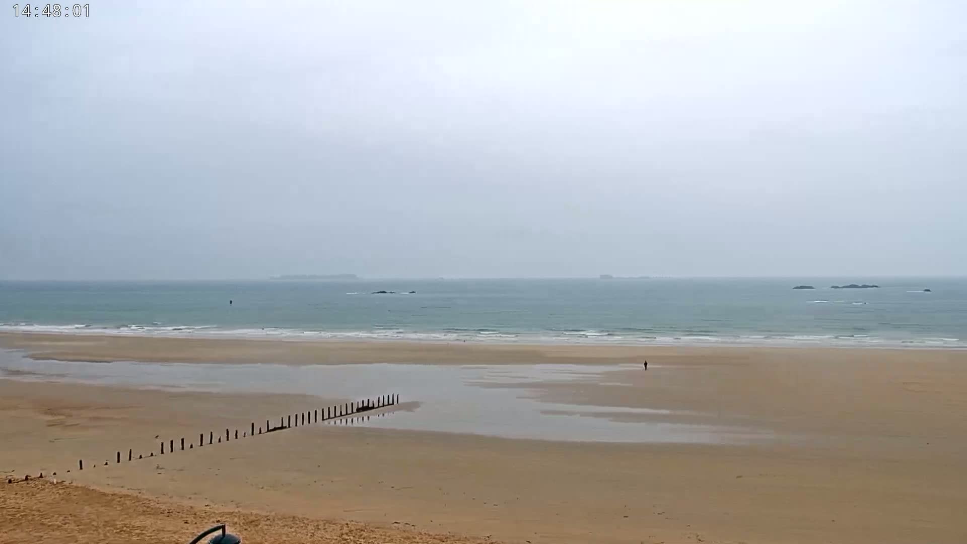 A sandy beach stretches to a calm, gray-toned sea under an overcast sky, featuring a low wooden structure on the sand and a lone figure walking in the distance.