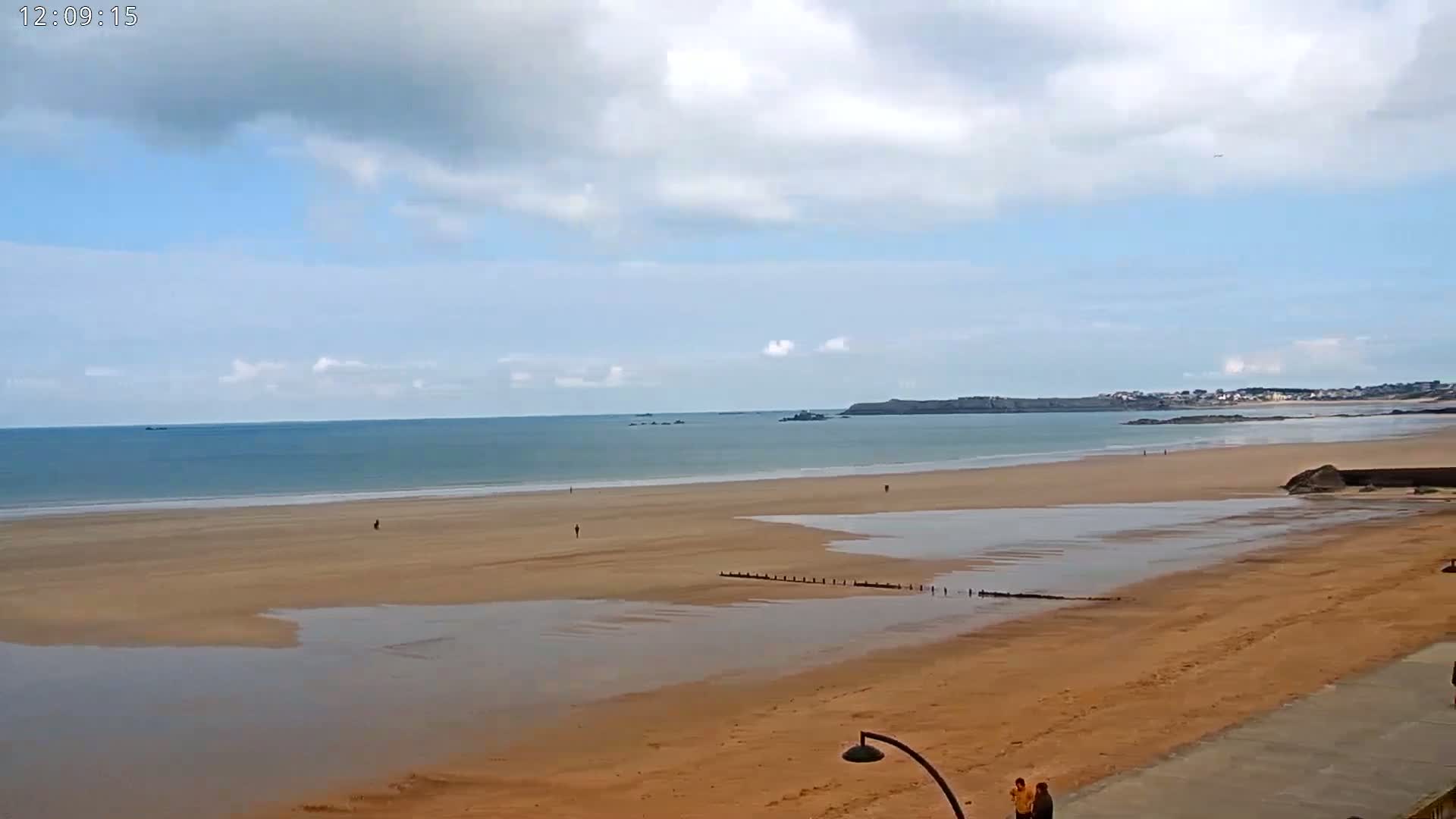 A wide sandy beach with calm ocean waters under a partly cloudy sky shows a few people walking on the sand.