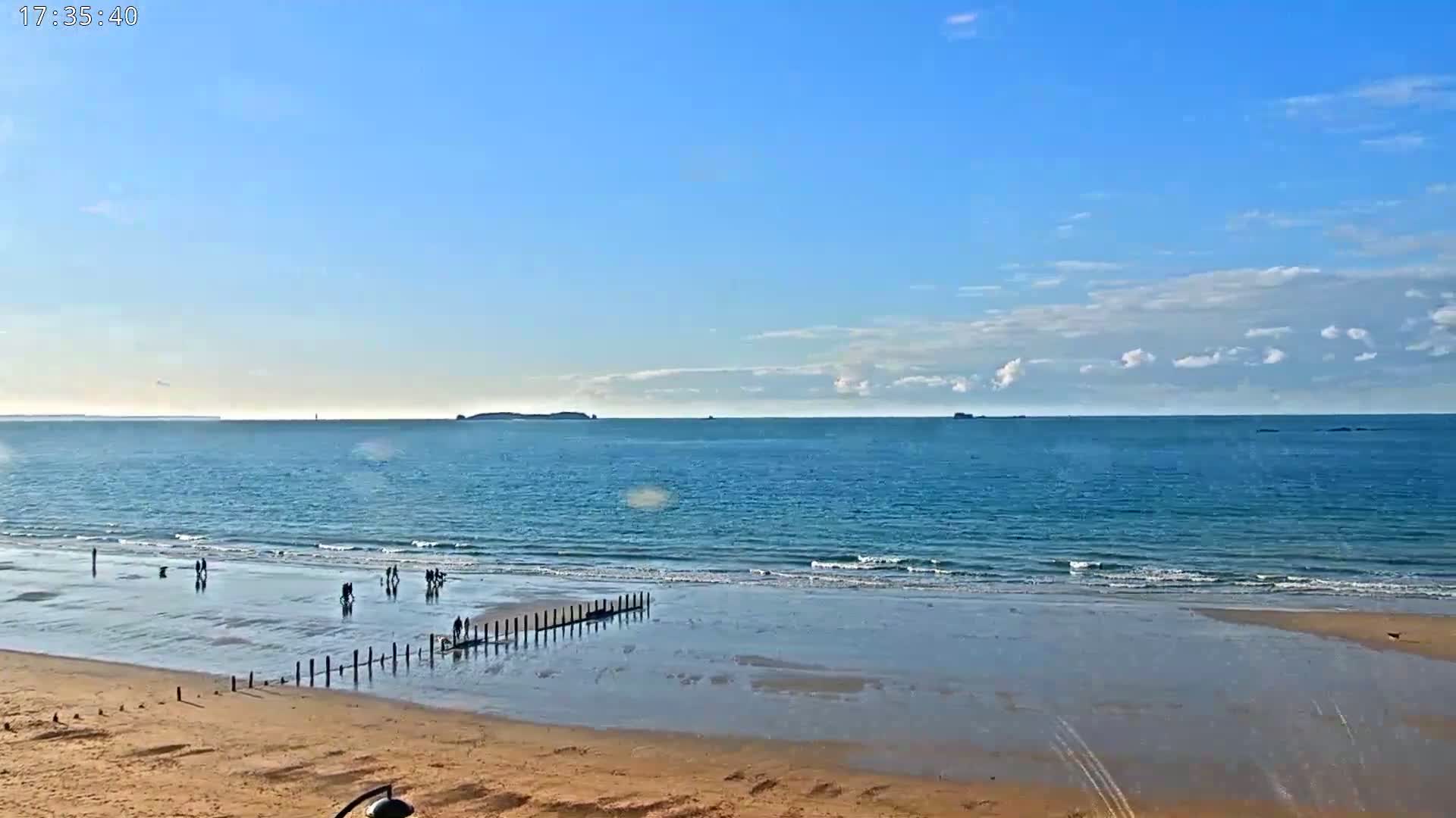 A sandy beach stretches out to a calm, blue ocean under a mostly clear sky with scattered clouds.