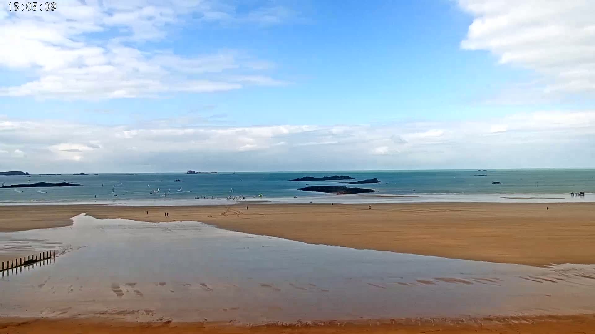 A mostly sunny day at a sandy beach shows many windsurfers on the water and a few people walking on the sand.